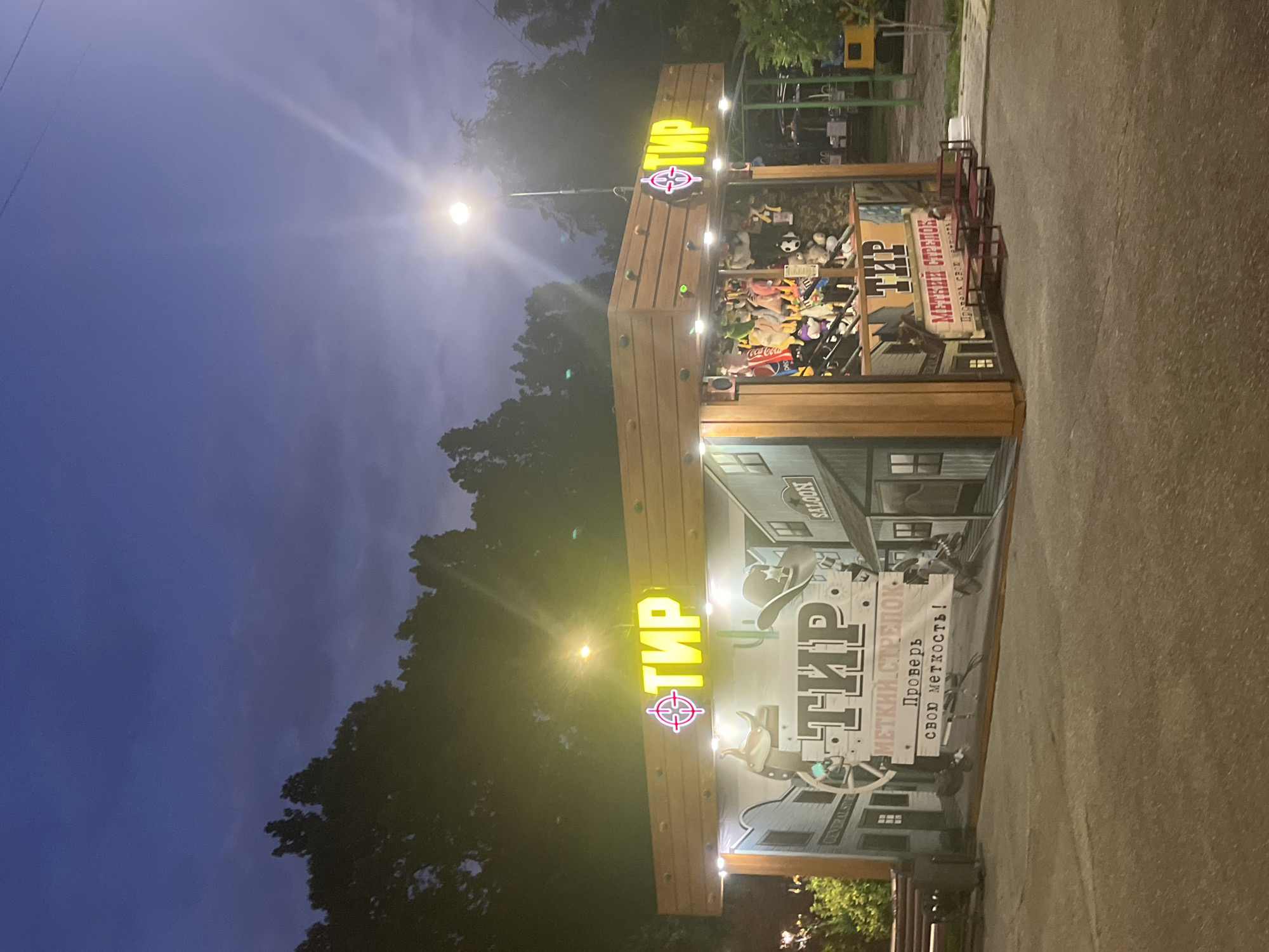 The image shows a shooting gallery attraction in a park in Krasnodar, Russia, at dusk. The attraction is designed with a Wild West theme, featuring painted murals of a town with a saloon and a general store. Above the attraction, a neon sign reads "ТИР" (Tir), which means shooting gallery. Below this sign, another sign reads "МЕТКИЙ СТРЕЛОК" (Metkiy strelok), meaning sharp shooter, and "Проверь свою меткость!" (Prover' svoyu metkost'!), meaning "Test your accuracy!". The shooting gallery itself is filled with stuffed toys that can be won. In the foreground, there is a paved area, and in the background, there are trees and a street lamp. No people are visible in the image.