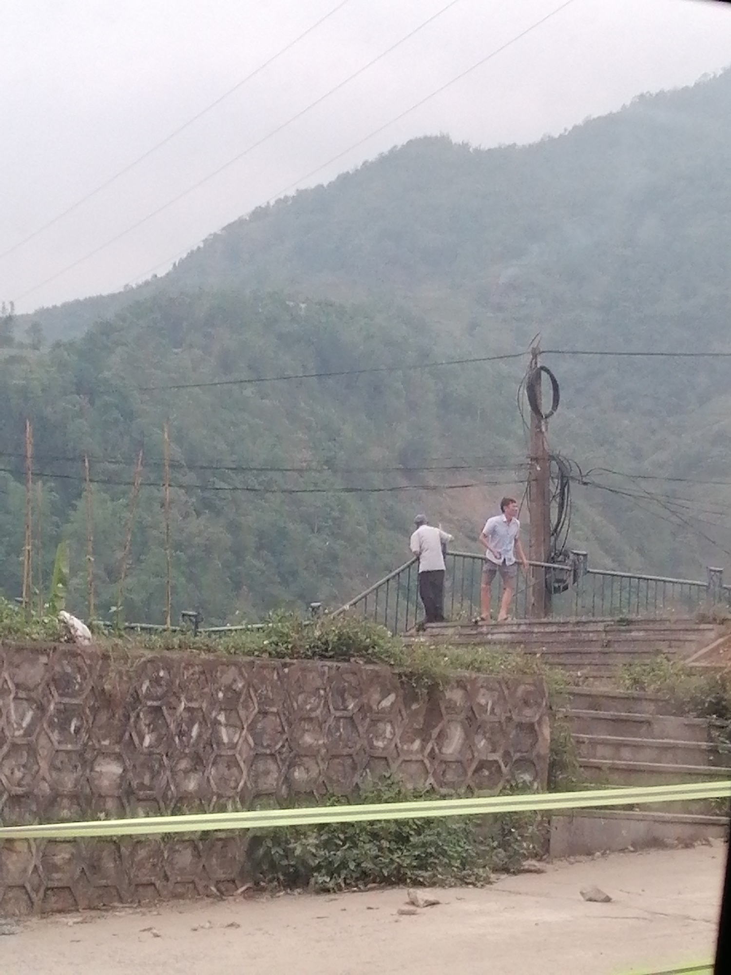 The image depicts a hazy, mountainous landscape in Trung Chải, Vietnam. In the foreground is a concrete retaining wall with a honeycomb pattern, partially obscured by vegetation and a horizontal green stripe. Above the wall is a set of concrete steps leading to a viewing platform with a dark metal railing.
Two people are visible on the platform. An older man in a light-colored shirt and dark pants leans against the railing, facing left. A younger man in a light-colored shirt and shorts stands next to a utility pole laden with wires, looking towards the landscape.
Behind the platform, a lush green hillside rises towards the top of the frame, fading into a misty sky. Several utility poles with power lines bisect the scene horizontally.