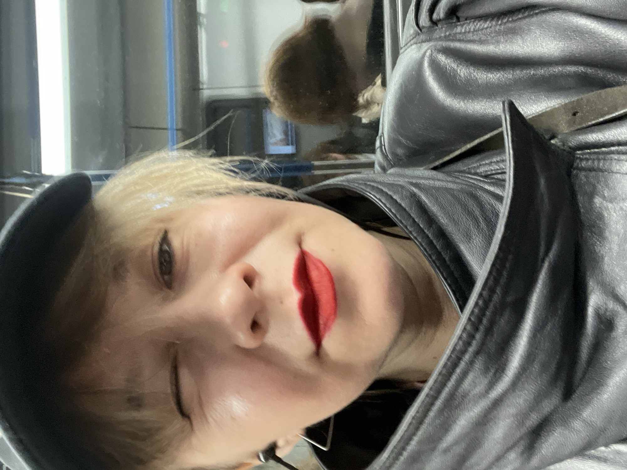 A woman with blonde hair and red lipstick is wearing a black leather jacket and a black cap. She is winking. Another person is visible in the background, reflected in a window. The location is likely a subway car in the city of Moscow, Russia, as suggested by the interior design elements and window.