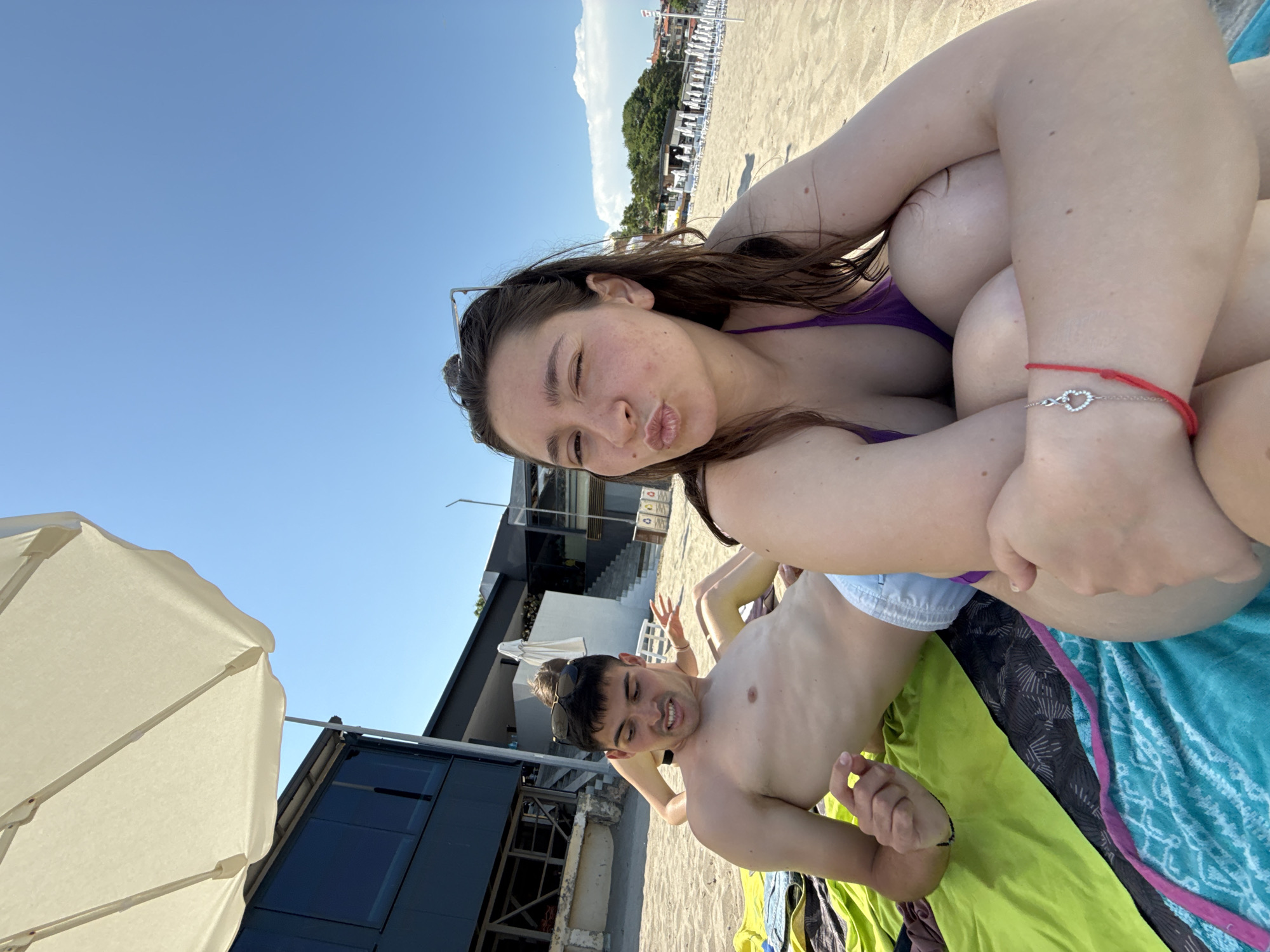 This casual, slightly tilted photo captures two young individuals relaxing on a sunny beach in Sozopol, Bulgaria. In the foreground, a young woman with long dark hair, wearing a purple bikini top and sunglasses pushed onto her head, looks towards the camera with a playful "kissing face" or pout. A red string bracelet and a silver chain bracelet with an infinity symbol and heart charm are visible on her wrist.
Beside her, a young man is lying down, shirtless and wearing light-colored swim shorts, smiling towards the camera. He also has sunglasses perched on his head. They are both resting on colorful beach towels; the woman appears to be on a lime green towel, and the man on a teal and white patterned towel.
The setting is a sandy beach under a clear, bright blue sky with a few white clouds. In the background, rows of white beach chairs and umbrellas line the shore. Behind these, there are buildings, including a modern structure with dark windows and stairs, likely part of a beach resort or restaurant. Lush green vegetation is also visible further back. A large, light-colored beach umbrella occupies the lower-left corner of the frame, providing shade. The time of day is clearly daytime, given the bright sunlight, suggesting a pleasant summer day. The overall atmosphere is relaxed and joyful. No visible text is present in the image.