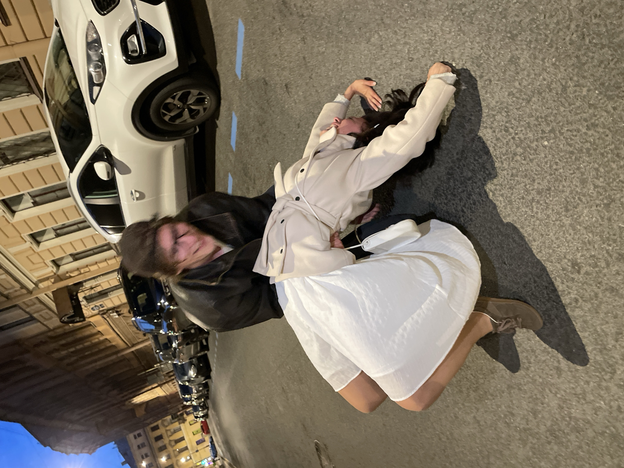 In Saint Petersburg, Russia, on a street at dusk, a person in a black leather jacket and glasses is leaning over to support another person who is bent backward in a dip on the asphalt. The person on the ground wears a white dress and a light-colored coat. Several cars are parked along the street, and buildings with windows line the background. The scene appears to be a playful or staged moment, possibly related to dancing or performance.