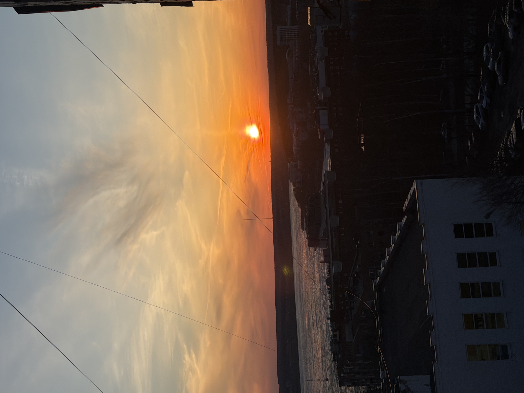 The image depicts a sunset over Izhevsk, Russia. The sun is positioned centrally, casting an orange and yellow glow across the sky. To the right, silhouetted buildings and structures are visible, including a large, white building with multiple windows. Below, a body of water reflects the light, with docks and small boats lining the edge. Power lines crisscross the sky in the foreground. The image is taken from a high vantage point, possibly a building or elevated structure.