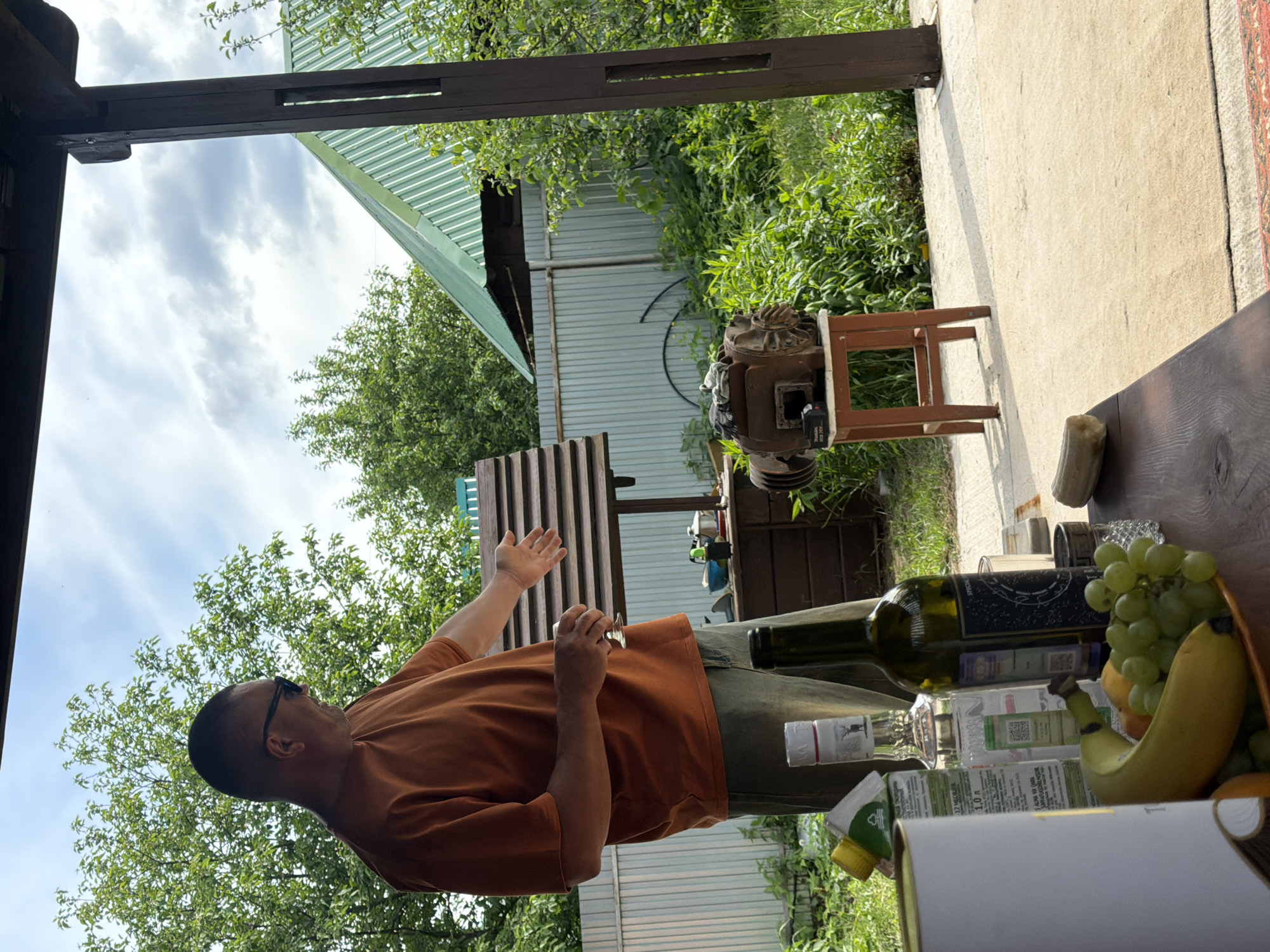 This image captures an outdoor scene, likely at a dacha or backyard in Chaikovsky, Russia, during the daytime under a partly cloudy sky.
The central figure is a man, viewed from the side, wearing a rust-colored short-sleeved t-shirt and olive green pants. He has short hair and wears sunglasses. His right hand is raised, palm open, in a gesturing or explanatory pose, while his left hand holds a small, light-colored object, possibly a utensil.
The man stands beside a dark wooden table that holds several items. Visible on the table are a dark green wine bottle, a clear spirit bottle (which displays "1.0 L" and some faint Cyrillic text, along with a QR code), and a white carton or bottle with a green cap, also marked "1.0 Л". A small bowl of fruit contains green grapes, a banana, and an orange. A light-colored food item, possibly a piece of bread, is also on the table.
To the right of the man, a heavy, weathered mechanical device, resembling an old engine part or industrial component, rests on a small wooden stool. The scene is framed by dark wooden beams of a pergola or canopy structure. The ground is a light-colored paved or concrete surface, with a corner of a patterned rug visible on the far right. In the background, there's lush green foliage from trees and bushes, a light-colored corrugated metal fence, and a building with a green corrugated metal roof. The overall atmosphere suggests a casual, relaxed outdoor gathering.
