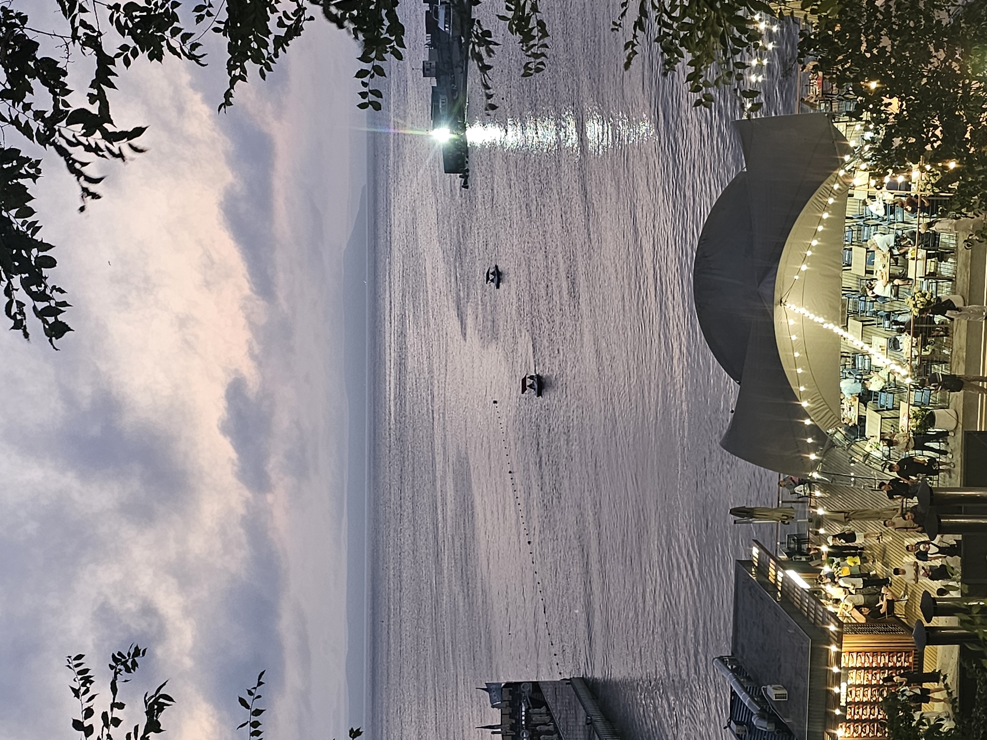 The image depicts a waterfront scene at dusk, likely in Vladivostok, Russia, given the style of buildings and the presence of boats. In the foreground, a busy outdoor seating area with tables and chairs is illuminated by strings of warm lights. People are seated at some of the tables, engaged in conversation or dining. Adjacent to this area is a building with windows displaying merchandise and a sign that appears to be in Cyrillic.
Beyond the seating area, a calm body of water stretches towards a distant, hazy coastline. Several small boats are visible on the water, one with an awning and passengers. In the mid-ground, a pier extends into the water, and further in the distance, buildings and structures line the shore, with a bright light source creating a reflection on the water's surface. The sky is overcast with soft pastel colors, indicating either sunrise or sunset. Foliage from trees frames the top and right side of the image, adding a natural element to the urban waterfront setting.