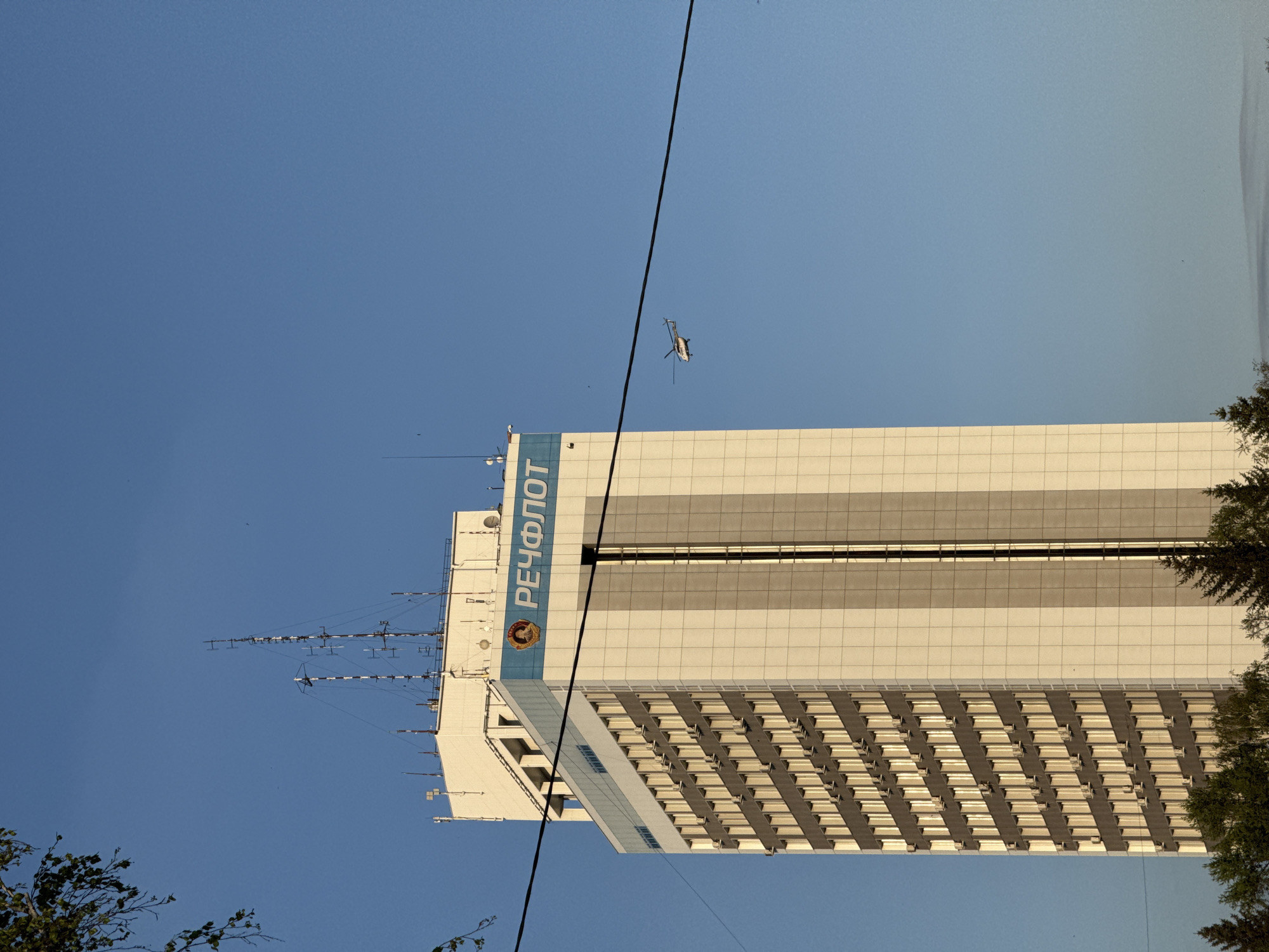 The image captures a low-angle view of the Rechflot building in Krasnoyarsk, Russia, against a clear blue sky. The building, a tall, rectangular structure, features a white façade with horizontal rows of recessed windows, creating a grid-like pattern. A section of the building has a blue stripe with the word "РЕЧФЛОТ" (Rechflot) vertically written in white Cyrillic characters. To the left of the writing is a circular logo. From the top of the building extends a network of radio antennae. A helicopter is seen flying in the sky near the building. A power line diagonally crosses the image. Trees frame the right and bottom left sides of the shot. The light indicates it is daytime.