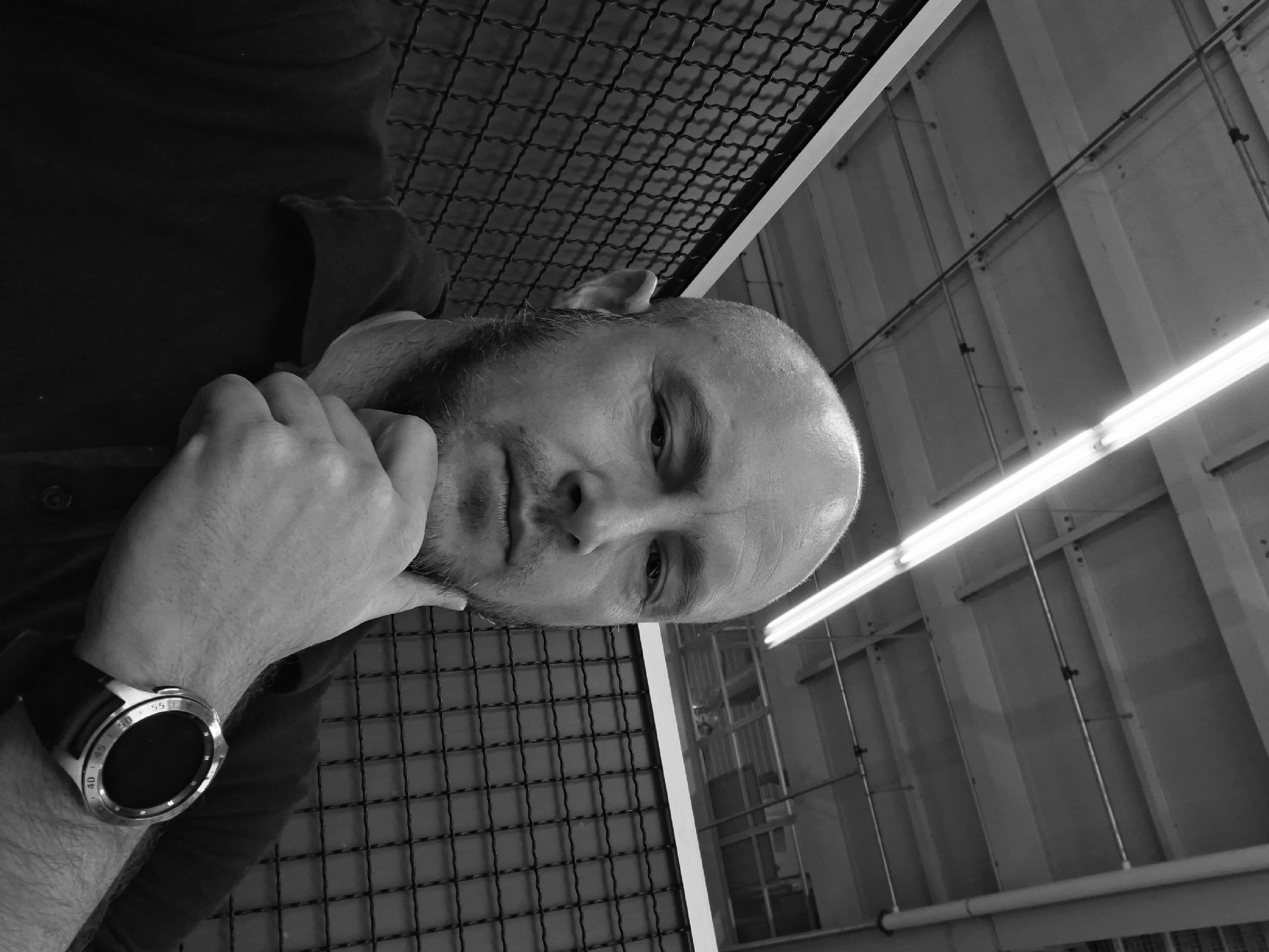 A black and white, medium shot, outdoor, waist-up, selfie, captured from a high angle, shows a bald man with a light beard and a dark polo shirt. He has his right hand resting on his chin and is wearing a digital watch on his left wrist. The man is situated in front of a black, wire mesh fence. Above him, fluorescent lights are visible, along with a ceiling with exposed conduits and beams. The background appears to be an industrial or warehouse-like setting.