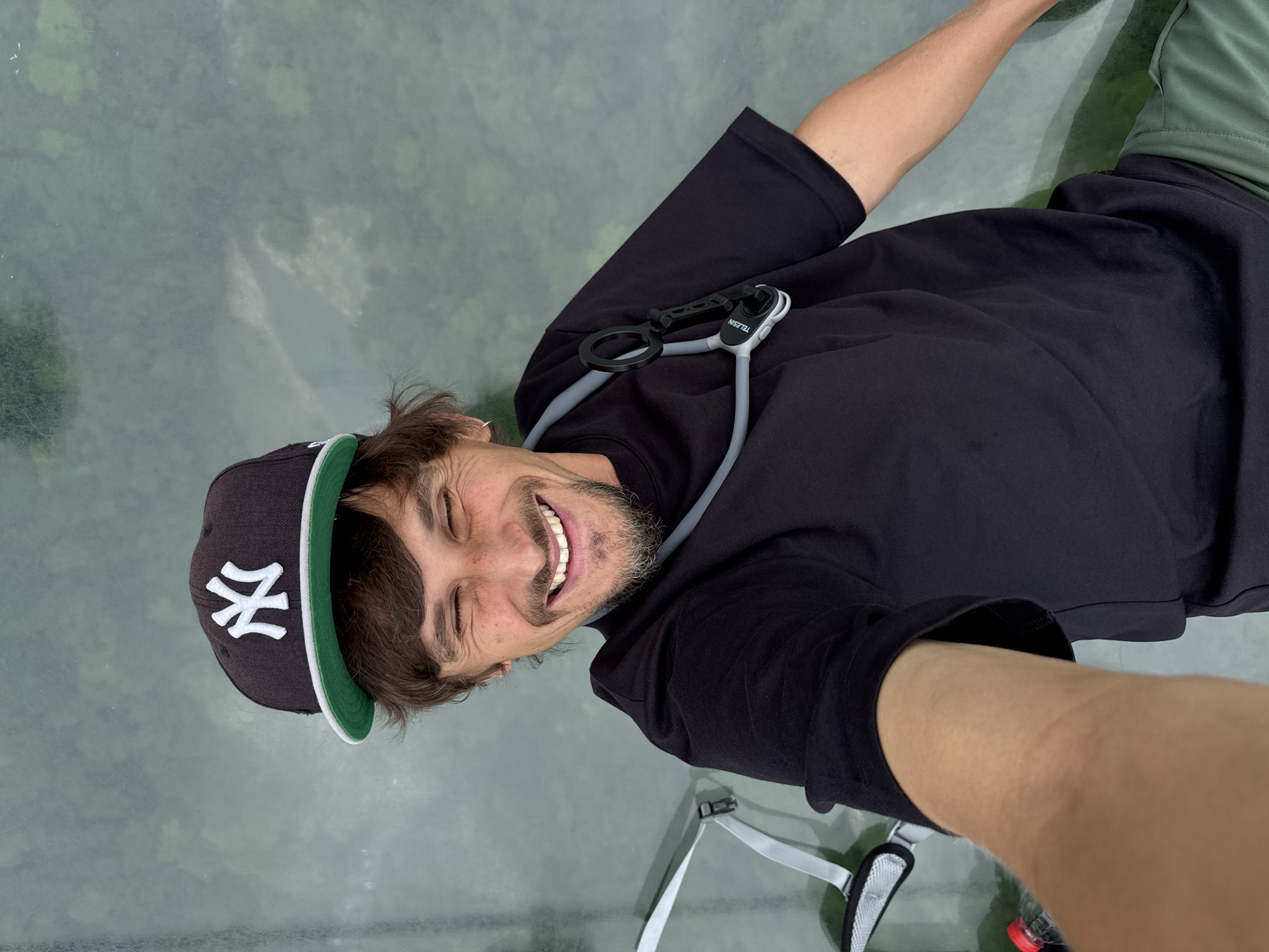 A man is smiling broadly while taking a selfie while standing on a glass bridge with a view of the forest below. He is wearing a black t-shirt, a New York Yankees baseball cap, and has a grey Teledsin neck mount around his neck. The background is a glass walkway above a forest in Zhangjiajie, China. In the foreground and bottom of the image, there is a partially visible white backpack strap, and a red water bottle cap.