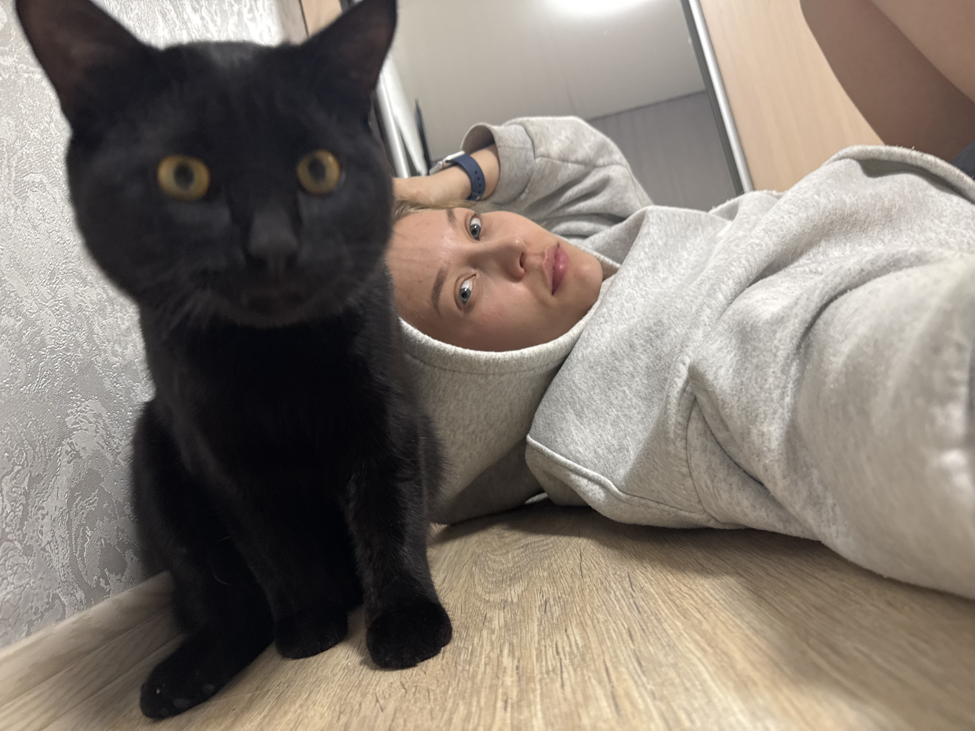 A young woman with light skin and blonde hair lies on a wooden floor, wearing a gray hooded sweatshirt and a blue wristband on her left arm. She looks directly into the camera. Next to her, slightly in front and to the left, a black cat with yellow eyes sits, also looking at the camera. The cat is positioned near a wall with patterned wallpaper. The wooden floor appears light in color. A door and part of a wall are visible in the background, implying an interior setting. The scene is casually lit, and the composition suggests a selfie or a spontaneous snapshot. Based on the information provided, this image was taken in the city of Chaikovsky, Russia.