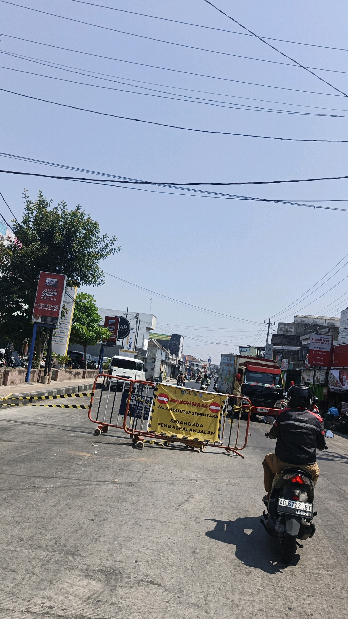 This image captures a street scene in Surakarta, Indonesia, on a clear, sunny day. In the foreground, a person rides a motorcycle, facing away from the camera. The motorcycle has a license plate reading "AD 6722 BY". The street is paved and appears to be a typical urban road with traffic.
Further down the road, a temporary road closure is in effect, marked by a red and yellow barricade. A yellow banner on the barricade displays text in Indonesian, including "MOHON MAAF JALANDITUTUP SEMENTARA" (Sorry, the road is temporarily closed) and "SEDANG ADA PENGASPALAN JALAN" (Road resurfacing is in progress). Beyond the barricade, various vehicles, including trucks and other motorcycles, are visible, suggesting ongoing traffic despite the closure.
The surrounding environment includes buildings with commercial signage, trees, and overhead power lines, characteristic of a bustling city street. The overall impression is one of everyday urban life disrupted by road maintenance.