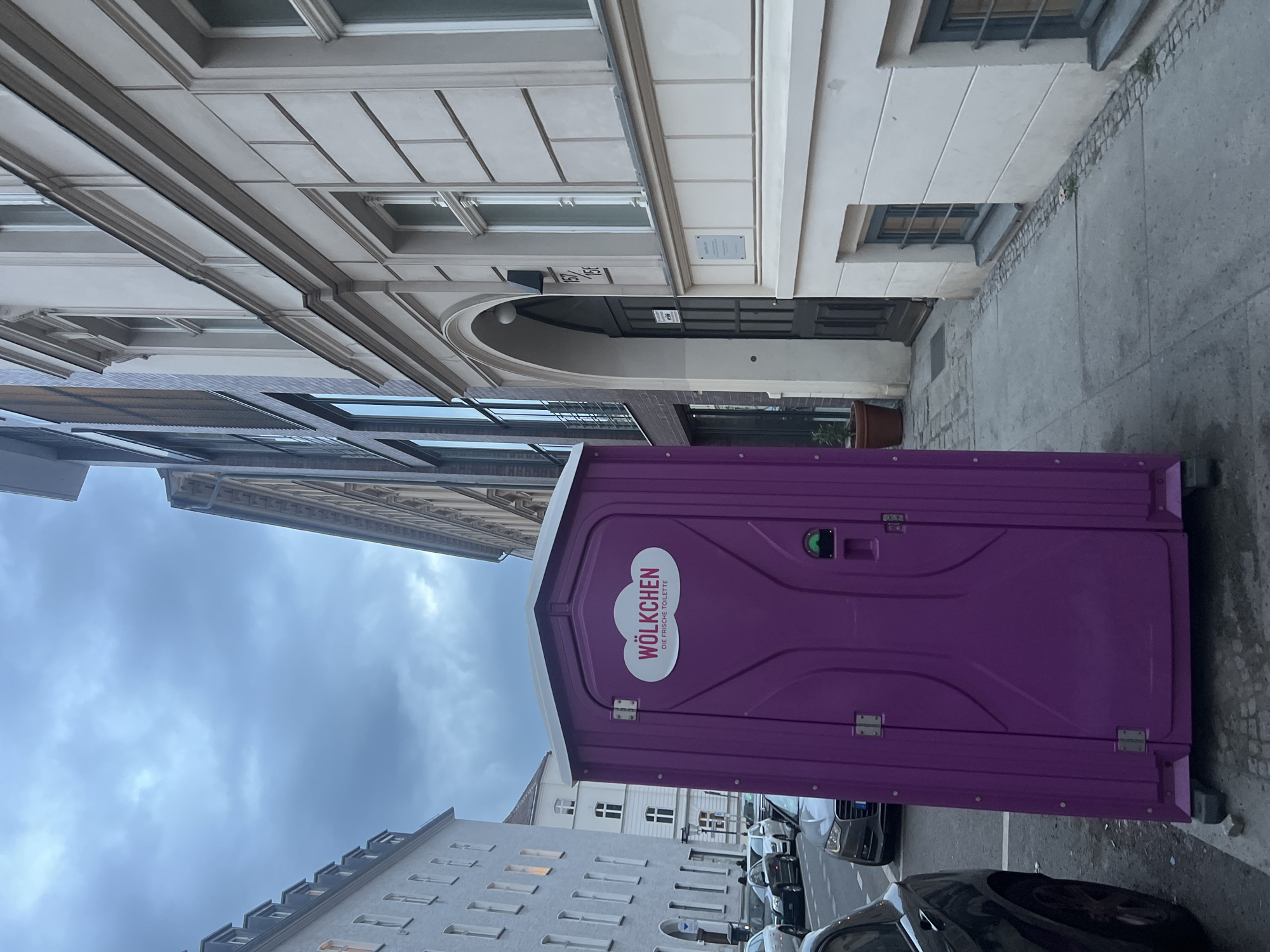 The image displays an urban street scene in Berlin, Germany, with a prominent purple portable toilet positioned horizontally across the lower central portion of the frame. The toilet's side, facing the viewer, features a white oval logo with "WÖLKCHEN" in bold red text and "DIE FRISCHE TOILETTE" in smaller black text below it. A green handle is visible on the door of the portable toilet.
Above the horizontally oriented portable toilet, extending upwards across the frame, are the facades of multi-story buildings. These buildings showcase typical European architecture with light-colored walls, numerous windows, and decorative cornices. One building features a dark arched entrance with the street numbers "154 / 156" clearly visible above it.
Below the portable toilet, more building facades are partially visible, along with a street where several dark-colored cars are parked. The ground to the right of the toilet is paved with a mix of concrete and cobblestones. The far left of the image is filled with an overcast and cloudy sky, indicating a cloudy day. No specific human activity is depicted; the scene captures a static moment in an urban environment.