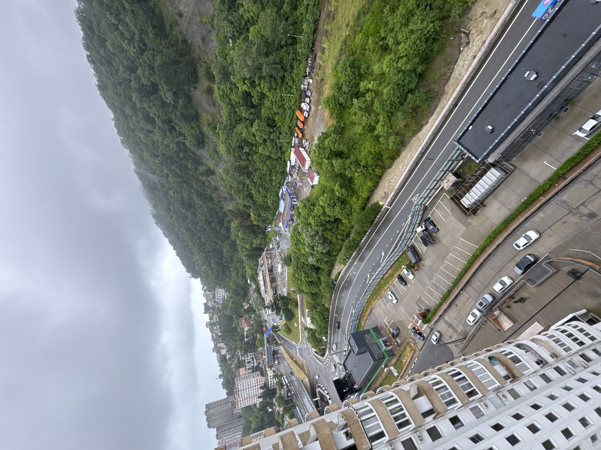 An aerial, high-angle, overcast, outdoor shot captures a city street scene with a steep, densely forested mountain rising in the background. The foreground features a curved, wet asphalt road with white lane markings and a pedestrian walkway separated by a metal railing. Several cars are parked in a lot next to the road, and a modern, multi-story building with a curved facade and many windows is partially visible on the left.
In the midground, the road continues, passing under an overpass with traffic. A small building with a green roof is situated near the road, and across from it, a row of service buildings, possibly a gas station and convenience stores, are visible with several vehicles parked in front. Orange buses are parked in a line to the right of the service buildings. The mountain, covered in lush green trees, dominates the upper right portion of the image, with a rocky incline visible in some areas. The sky is heavy with gray clouds, suggesting rainy weather. The overall impression is of a coastal city built against a natural, mountainous backdrop. The scene appears to be in Sochi, Russia, likely along a major thoroughfare connecting the coast to higher elevations.