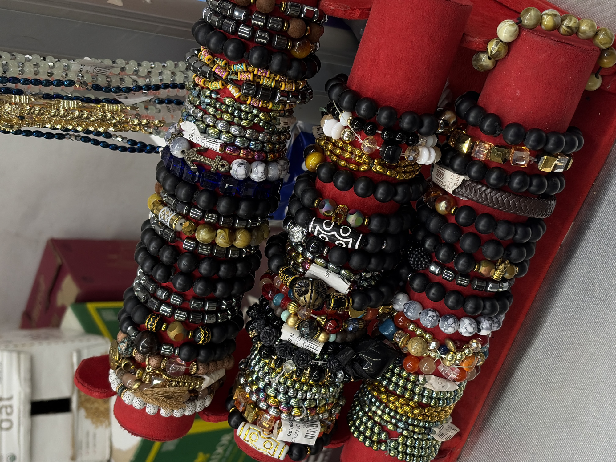 A close-up, eye-level shot displays an array of beaded bracelets arranged on red velvet display stands. The bracelets are predominantly black, with variations in bead size, texture, and accent colors such as gold, silver, and opaque white. Some bracelets feature charms, including a golden anchor, a golden coin, and decorative metal beads. In the background, blurred elements suggest a retail environment, with boxes labeled "oat" and other merchandise visible. The overall impression is that of a jewelry display, possibly in a market or shop. The image composition is focused on the bracelets, with the background intentionally out of focus.