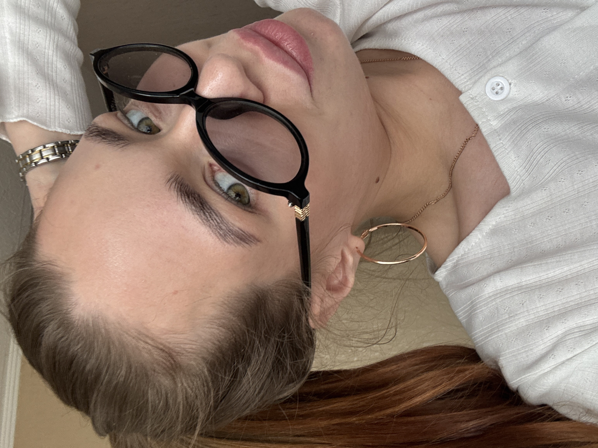 A woman is seen from a tilted, close-up perspective. She wears black framed sunglasses, a white textured shirt with a button, and a gold necklace and hoop earring. She also has a silver watch on her wrist. Her hair is brown with reddish undertones. The location in the city of Chaikovsky, Russia, is not identifiable from the provided image.