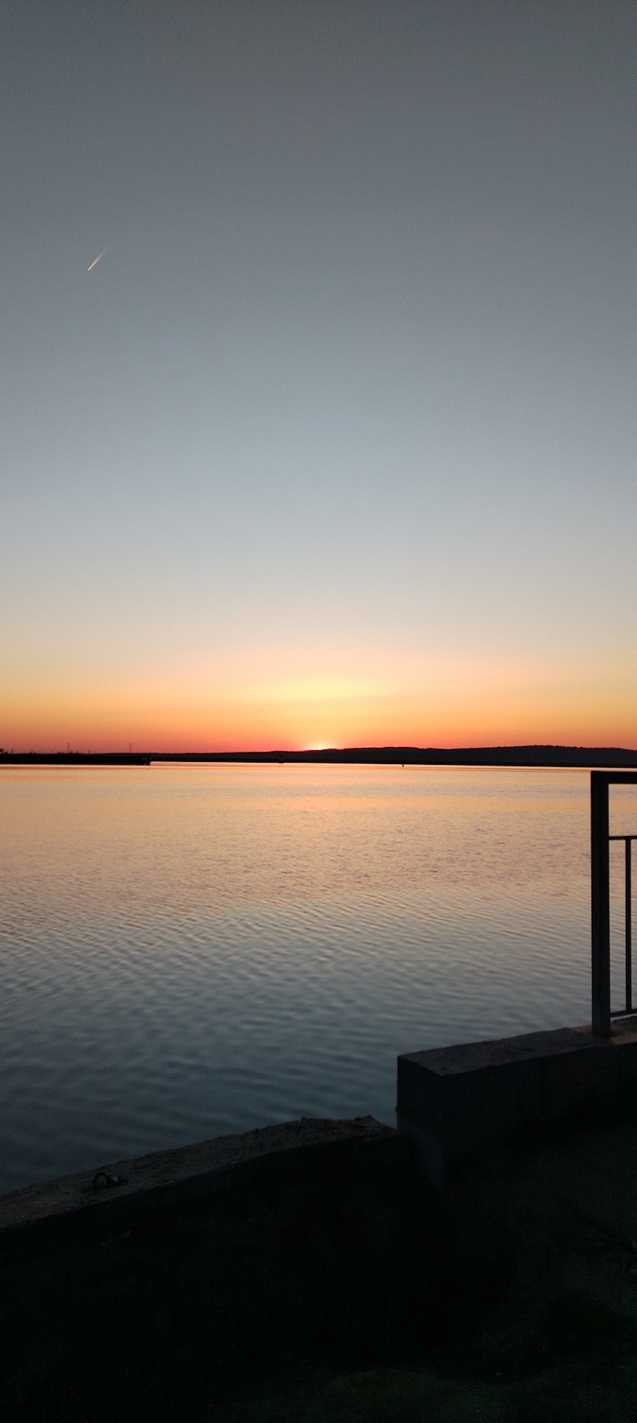 The image depicts a tranquil scene at sunset over a body of water, most likely a lake. A thin strip of land is visible in the distance, silhouetted against the horizon. The sky transitions from light blue at the top to a vibrant orange and red near the horizon where the sun is setting. The water surface reflects the colors of the sky, with gentle ripples creating a sense of calmness. In the foreground, a section of a pier or embankment is visible, with a railing suggesting a viewing point. The location is Unknown, Unknown.