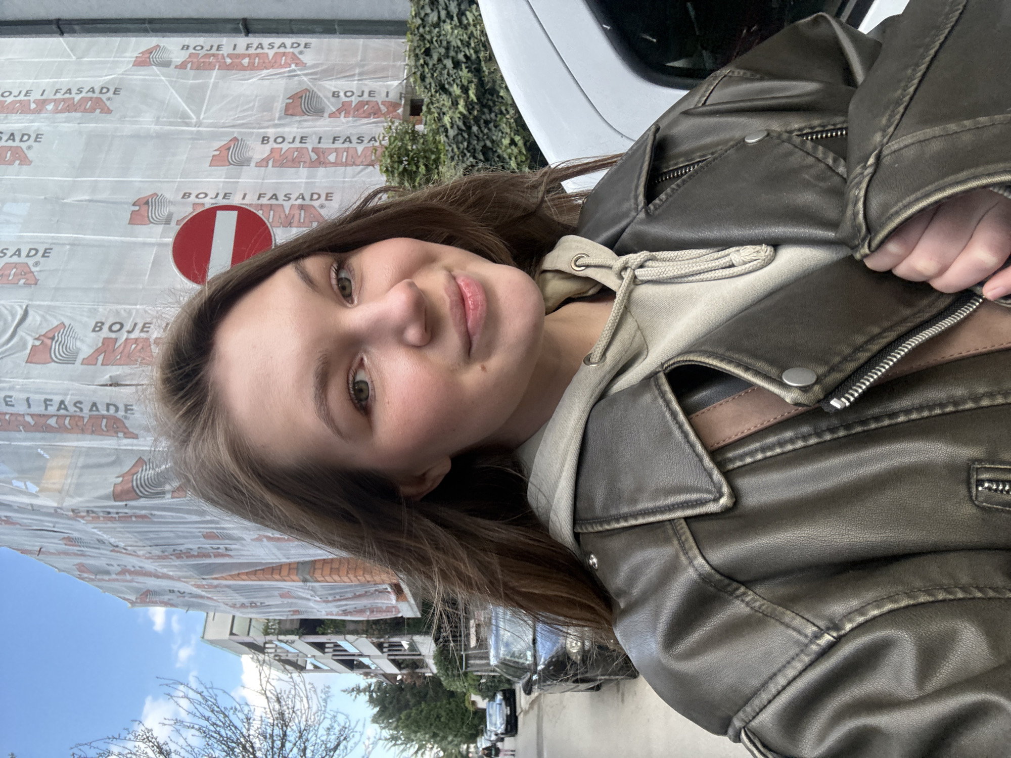 Here is a forensic description of the image:
The image is a selfie of a young woman in an urban environment. She is wearing a leather jacket over a hooded sweatshirt. In the background, there are buildings and a construction site covered in tarpaulins with the text "BOJE I FASADE MAXIMA". There is also a "no entry" sign visible. Trees line the street and a few cars are parked along the road. Based on the building style and the signage, the location is in Vozdovac Urban Municipality, Serbia.