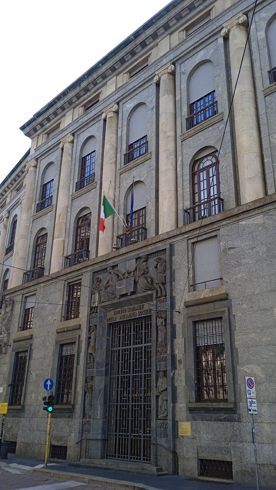 The image shows the exterior of a large, imposing stone building, likely a public institution, in Milan, Italy. The building features classical architectural elements such as large columns and arched windows with ornate detailing. The facade is made of grey stone, with some sections appearing to be polished granite.
Above a grand entrance gate, there is a large bas-relief carving depicting several figures and what appears to be an inscription in Italian. The inscription reads "ISTITUTO NAZIONALE DELLA PREVIDENZA SOCIALE" which translates to "National Institute of Social Security." This confirms the building is the INPS (Istituto Nazionale della Previdenza Sociale) headquarters.
Two flags are visible on balconies above the entrance: the Italian flag (green, white, and red) and another flag, possibly blue. There are several windows on multiple floors, some with closed shutters and others with metal grates.
In the foreground, a street scene is depicted. A yellow lamppost with a traffic light shows a green light. A blue circular traffic sign with a white arrow indicates "straight ahead." A smaller yellow sign is visible on the lamppost. A trash bin is located near the base of the lamppost. On the right side of the image, a parking sign with several indications is mounted on a pole. The ground is paved with stone. The sky above is overcast.