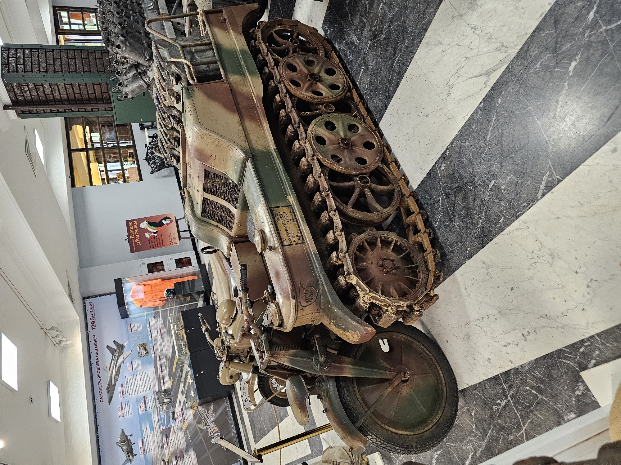 The image captures an exhibit featuring a historical vehicle with tank treads and a single front wheel, displayed indoors on a black and white striped floor. The vehicle, painted in camouflage colors, is positioned near an exhibit wall showcasing aviation history. A sign displaying the names of "Yakovlev" aircraft is prominently visible. The exhibit is located in a museum or historical center with an area about airplanes and Яковлева over the sea, possibly in Архангельское, Russia. An informational poster featuring an advertisement is visible in the background along with displays behind glass.
