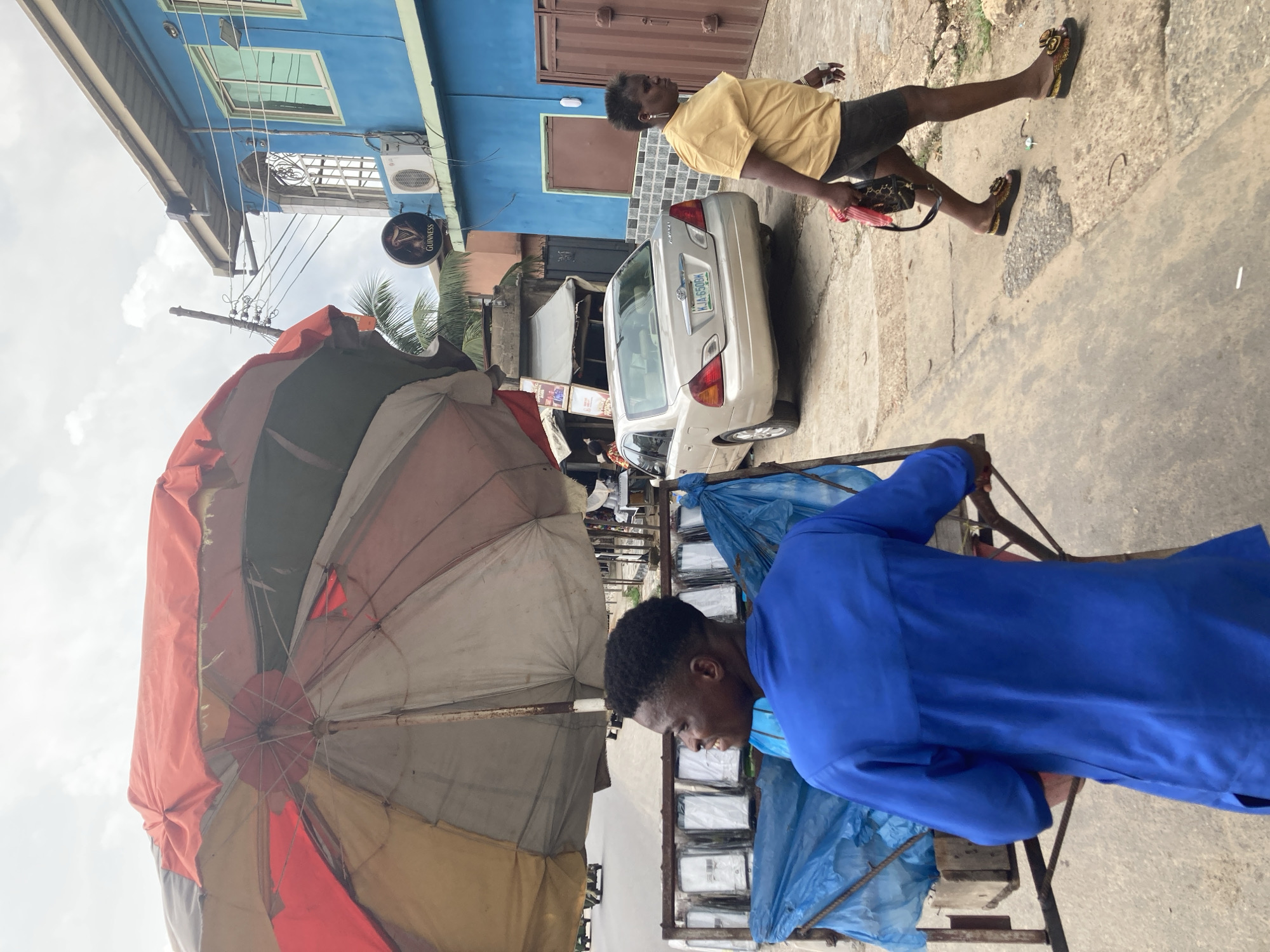 The image shows a street scene in Aba, Nigeria. A man in a blue shirt is pulling a cart with packaged goods under a large, multi-colored umbrella. A woman in a yellow shirt and black shorts is walking in the background. A white car with the license plate KJA-650BM is parked nearby. A Guinness sign is visible on a blue building. The environment appears urban and somewhat cluttered.