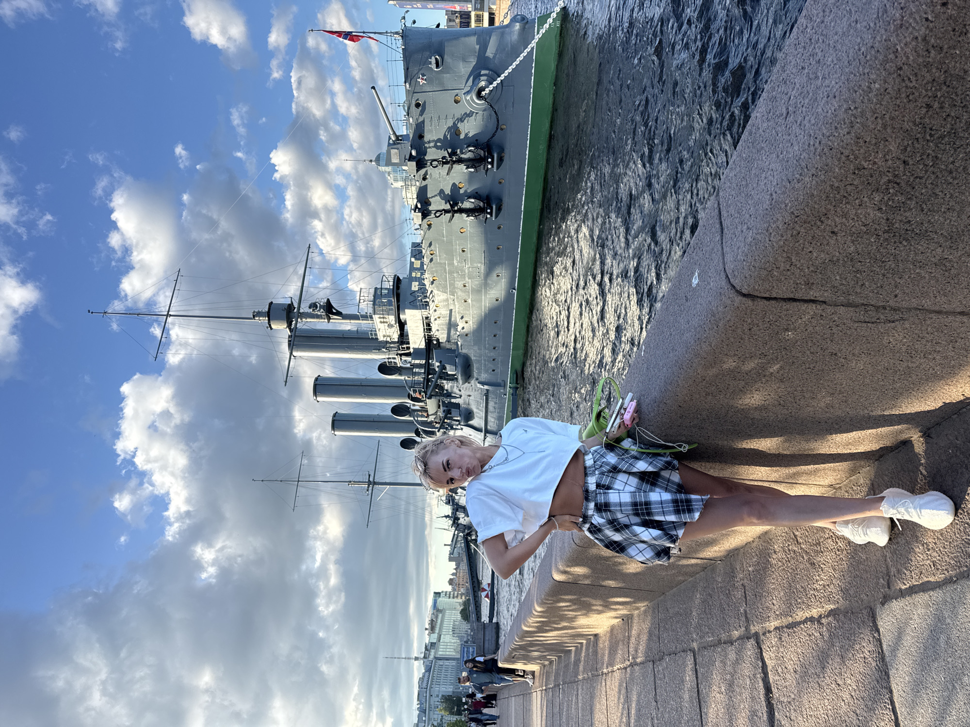 A blonde woman with sunglasses perched on her head poses in front of a historic warship docked on the Neva River in Saint Petersburg, Russia. She wears a white crop top, a black and white plaid skirt, and white sneakers, with a green bag and a pink object in her hands. Behind her, the Aurora cruiser, a prominent museum ship and symbol of the Russian Revolution, is moored. The ship is a grey, four-masted vessel with visible cannons and anchors. In the background, the cityscape of Saint Petersburg unfolds under a sky filled with dramatic clouds. The woman stands on a stone embankment, which lines the river and separates the walkway from the water. The lighting suggests it is late afternoon, with the sun casting shadows and highlighting the textures of the stone and the water.