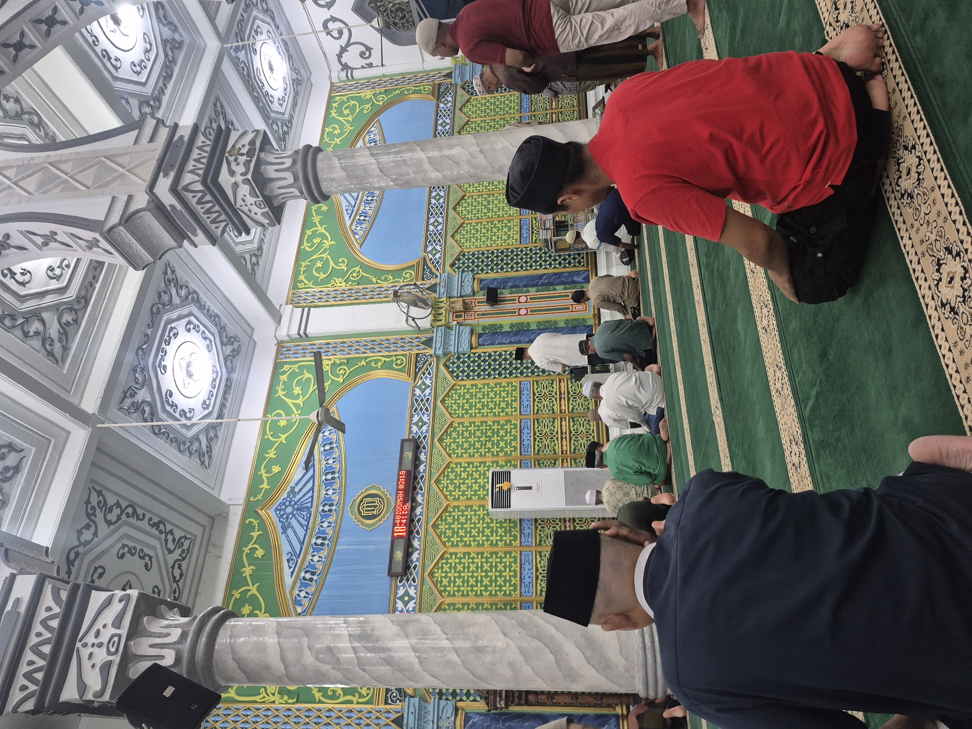 The image captures a group of Muslim men in Keudekruenggeukueh, Indonesia, engaged in prayer inside a mosque. They are kneeling on a green carpet, aligned in rows, facing a wall adorned with colorful, geometrically patterned decorations. A digital clock is mounted on the wall displaying what appears to be prayer times. The mosque's interior is characterized by ornate columns and detailed ceiling designs. The men are wearing various attires, including shirts and head coverings, and appear to be focused on their prayer.
