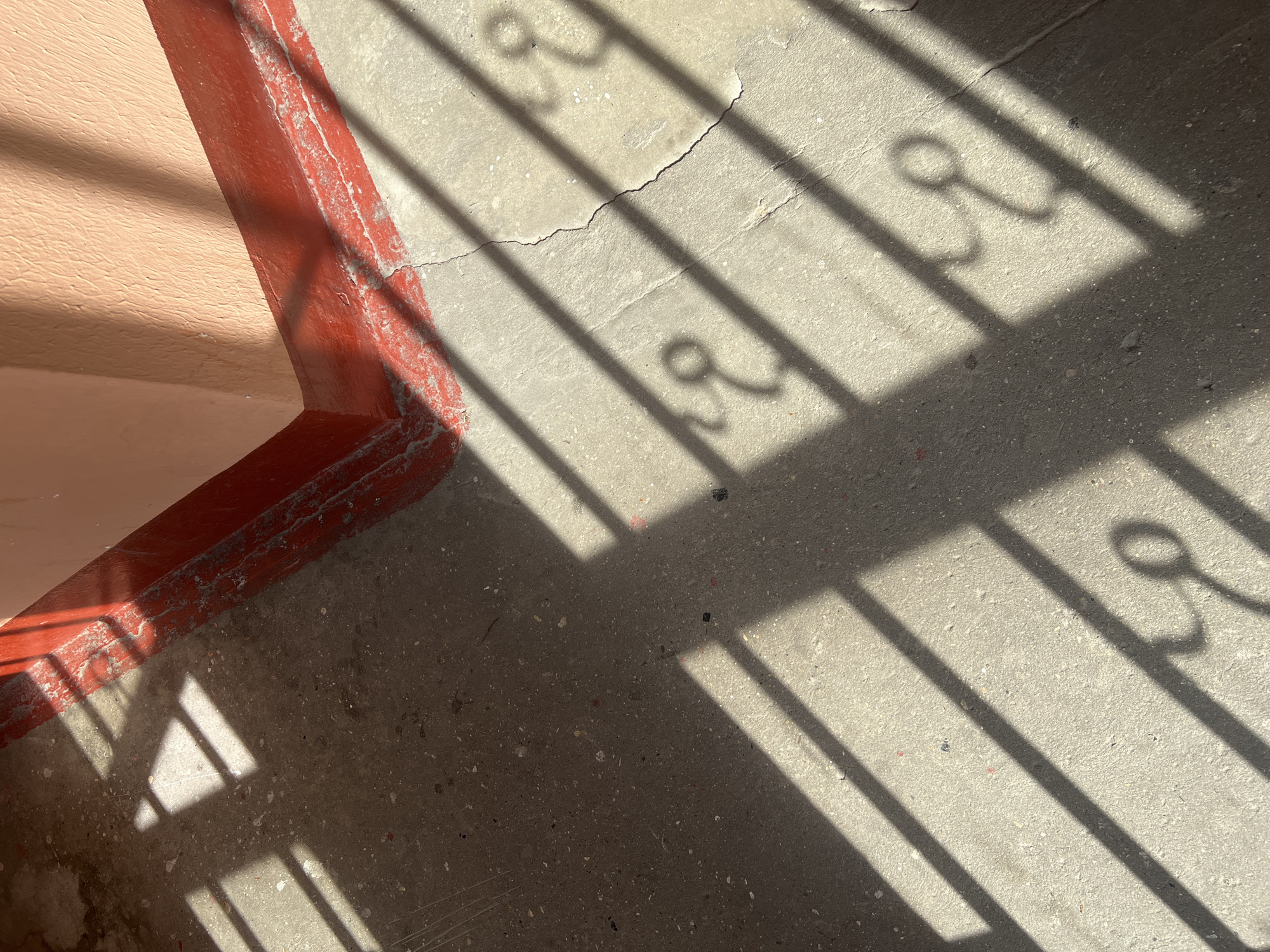 The image depicts a close-up of a corner where a peach-colored wall meets a gray concrete floor. The edge of the wall is marked with a thick, red border. Shadow patterns, likely from a barred window or gate, are cast across the floor, featuring straight lines and distinctive curled elements. The floor shows minor surface imperfections and a crack. The location cannot be determined from the image.