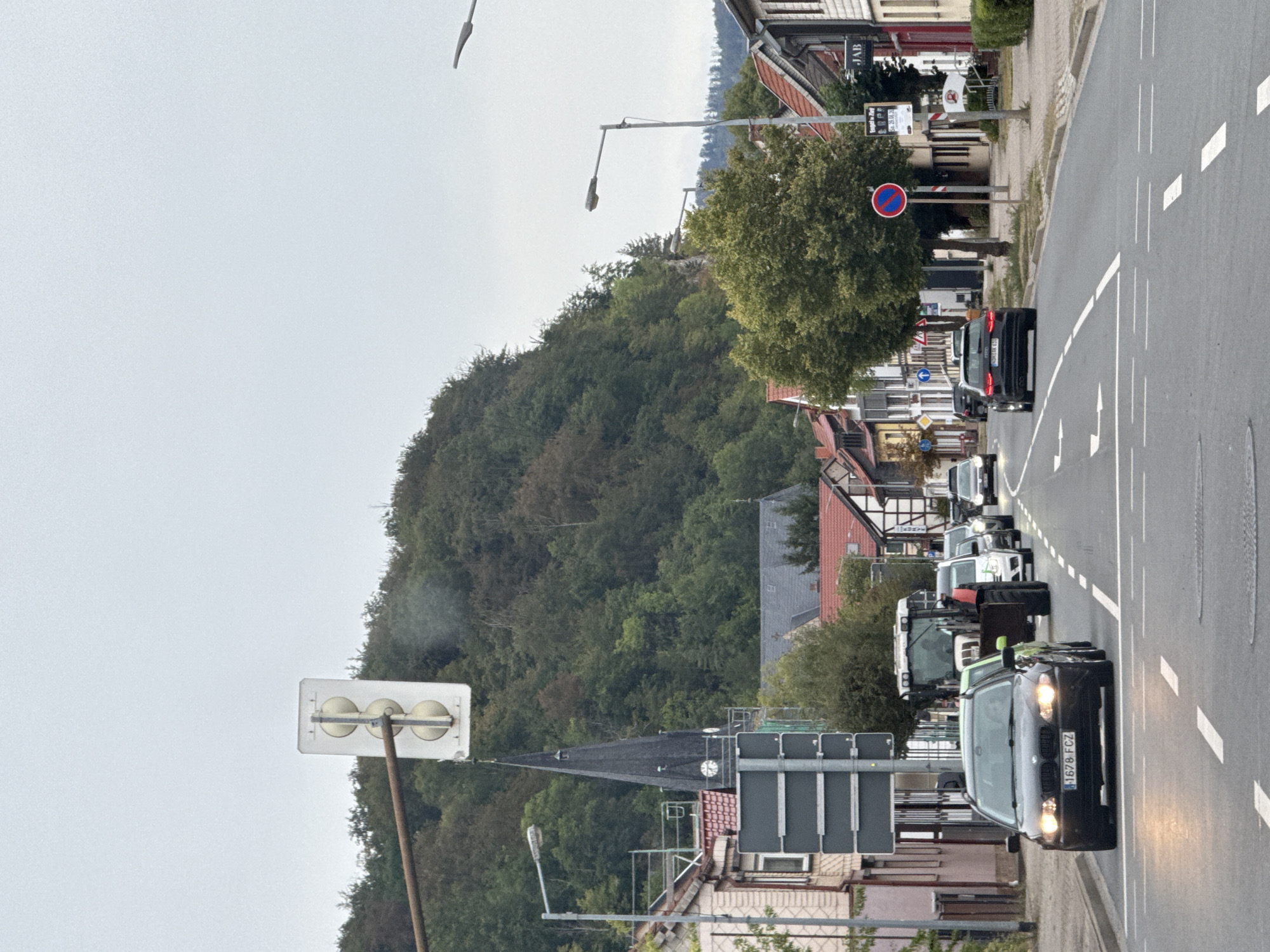 The image shows a street scene in Niedersachswerfen, Germany, with a slight incline. On the left side of the street, there are several buildings, including a church with a tall, dark steeple. A traffic light is visible overhead. The street is lined with trees and more buildings on the right. Several vehicles are on the road, including a black BMW in the foreground, a white tractor, and other cars behind it. The sky is overcast.