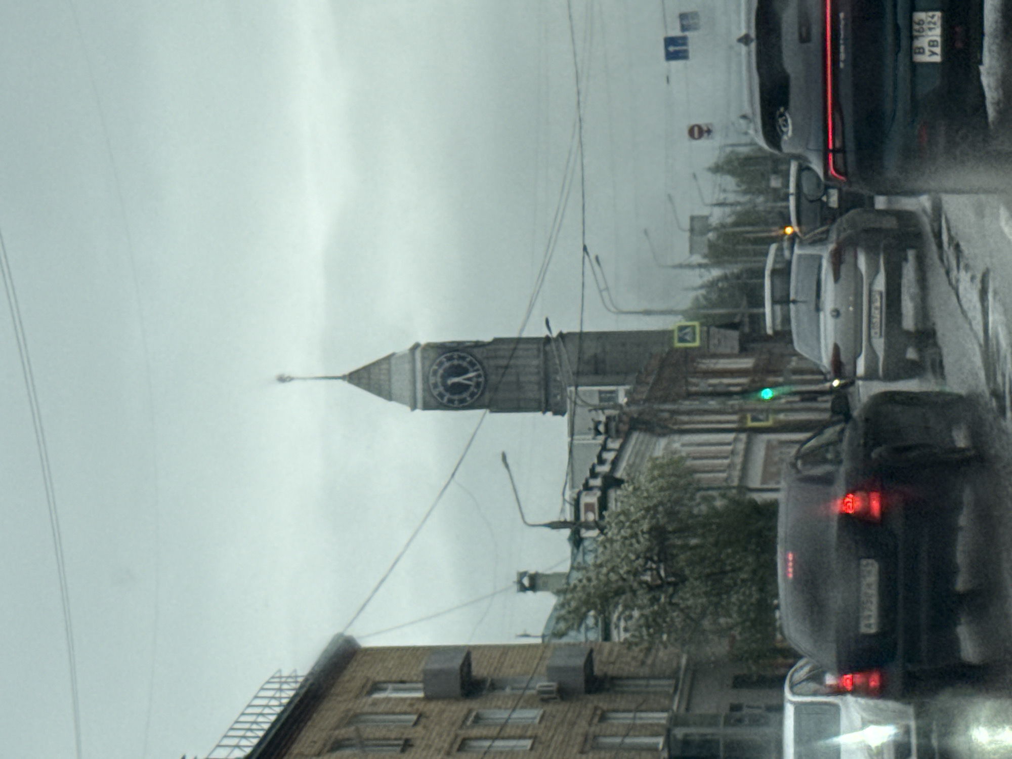 The image depicts a cityscape of Krasnoyarsk, Russia, on a cloudy day, possibly raining. A prominent feature is a clock tower, likely the Krasnoyarsk Clock Tower, rising above the buildings. The scene is viewed from a moving vehicle on a wet road, with other cars visible in the foreground. The traffic signal appears to be green. Buildings of varying heights and styles are present, and utility wires are strung across the sky. The overall atmosphere is urban and somewhat gloomy.