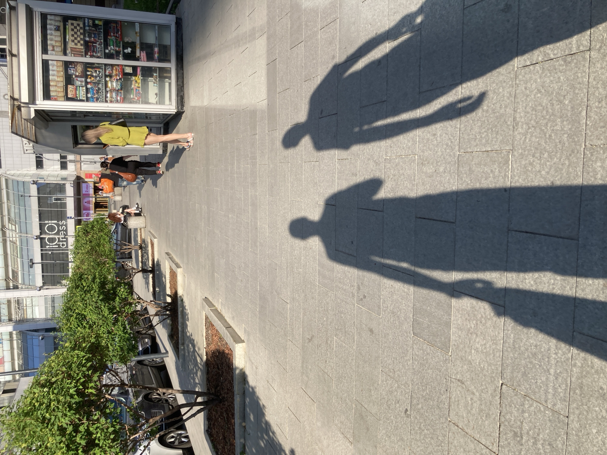 The image shows a pedestrian walkway paved with gray rectangular stones, likely in a city environment. On the left, there is a newsstand with displayed items, and a woman with blonde hair, wearing a yellow outfit and sandals, is standing near it. Further along the walkway, a few other people are visible. A row of small trees planted in rectangular beds lines the left side of the walkway, and buildings are visible in the background. Shadows of two people are cast onto the walkway, indicating a sunny day. The location is likely Moscow, Russia based on the newsstand architecture and overall urban appearance.