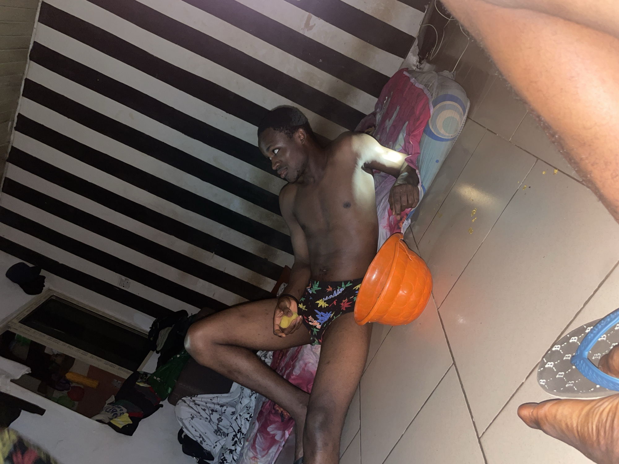 The image shows a young Black man lying on a bed in what appears to be a bedroom in Ihiagwa, Nigeria. He is shirtless and wearing patterned underwear. He is holding a yellow object in his right hand and an orange plastic bowl is to his right. The room has tiled flooring and a wall with diagonal black and white stripes. Several pieces of clothing are scattered around the room, including on the bed and near a window. A blue flip-flop is visible on the floor in the foreground. The lighting suggests it is nighttime or indoors with artificial light. There is no clear indication of the activity or event taking place, but the man's posture and the overall scene suggest a state of rest or possibly unease.