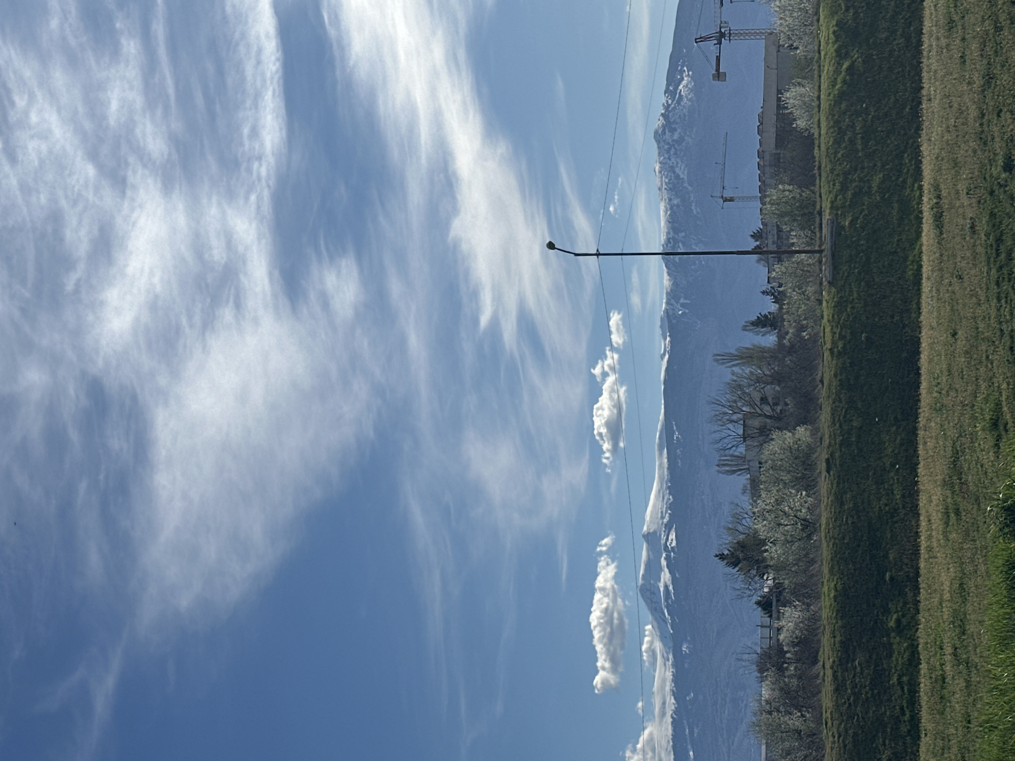 The image depicts a scene in what appears to be an urban-adjacent area, with a view encompassing sky, mountains, and a grassy verge. The sky is mostly clear blue, filled with cirrus and cumulus clouds. In the distance are mountains with snow on their peaks. A line of trees runs along the middle ground. Construction cranes are also visible, suggesting ongoing development. A single street lamp is near the center of the frame.
The location is likely in or around Sofia, Bulgaria, considering the mountain range is probably the Vitosha mountain or the Balkan Mountains, which are prominent features of the Sofia landscape.