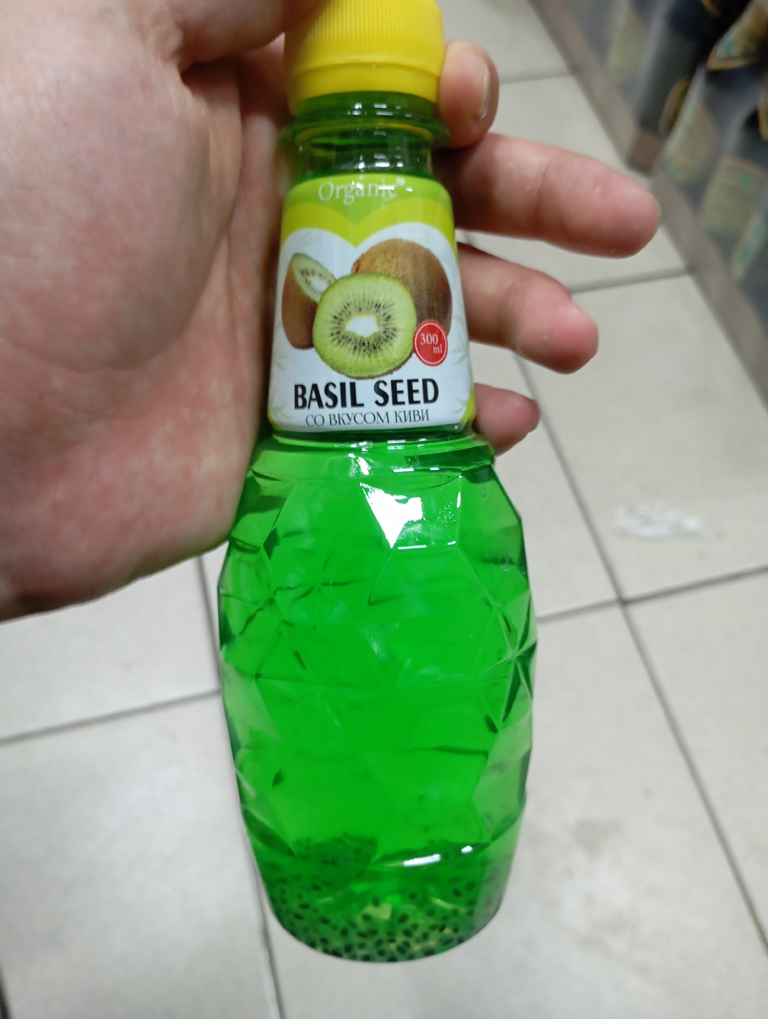 A hand is holding a green plastic bottle of "Basil Seed" drink with kiwi flavor. The bottle has a yellow cap and a label with images of kiwi fruit. The drink inside is green and contains many small black seeds at the bottom. The bottle is held in front of a tiled floor, possibly in a retail environment given the blurry background of shelves with bottles. The lighting is artificial, casting some shadows. The image was taken in Moscow, Russia.