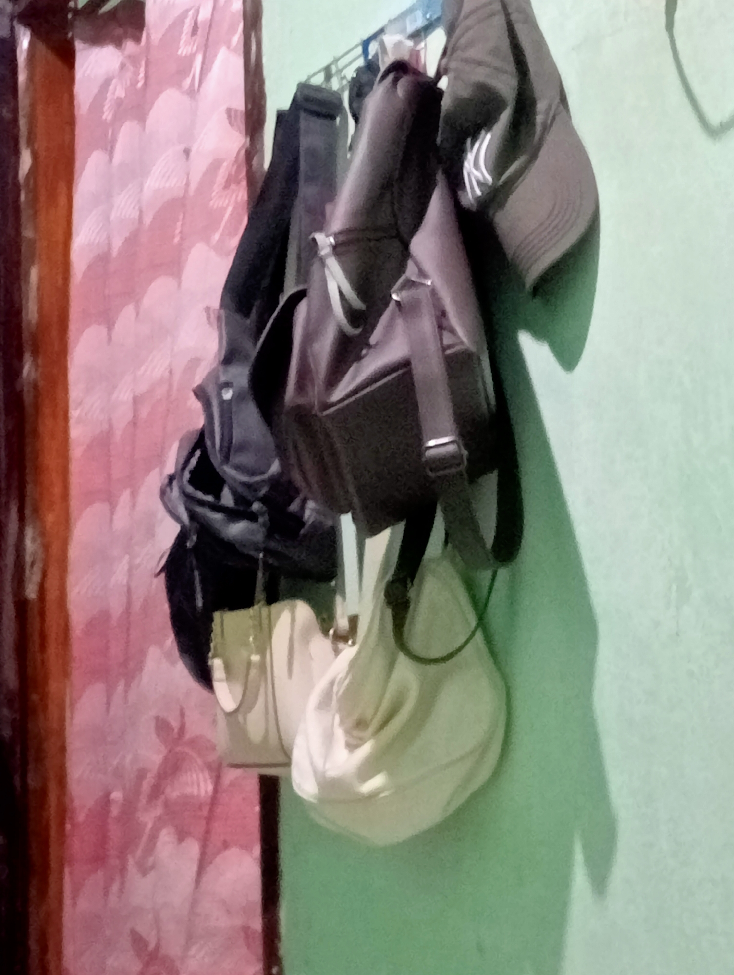 The image shows several bags and a baseball cap hanging on a rack against a pale green wall. The bags appear to be of various sizes and colors, including black and off-white. The baseball cap is dark gray with a white logo. To the left of the bags and wall is a curtain with a pink and white patterned design, and a dark vertical element that may be a doorframe or furniture. The lighting suggests an indoor environment, possibly a residential space. The scene is located in Kota Negara Timur, Indonesia, where such arrangements of personal belongings are common.