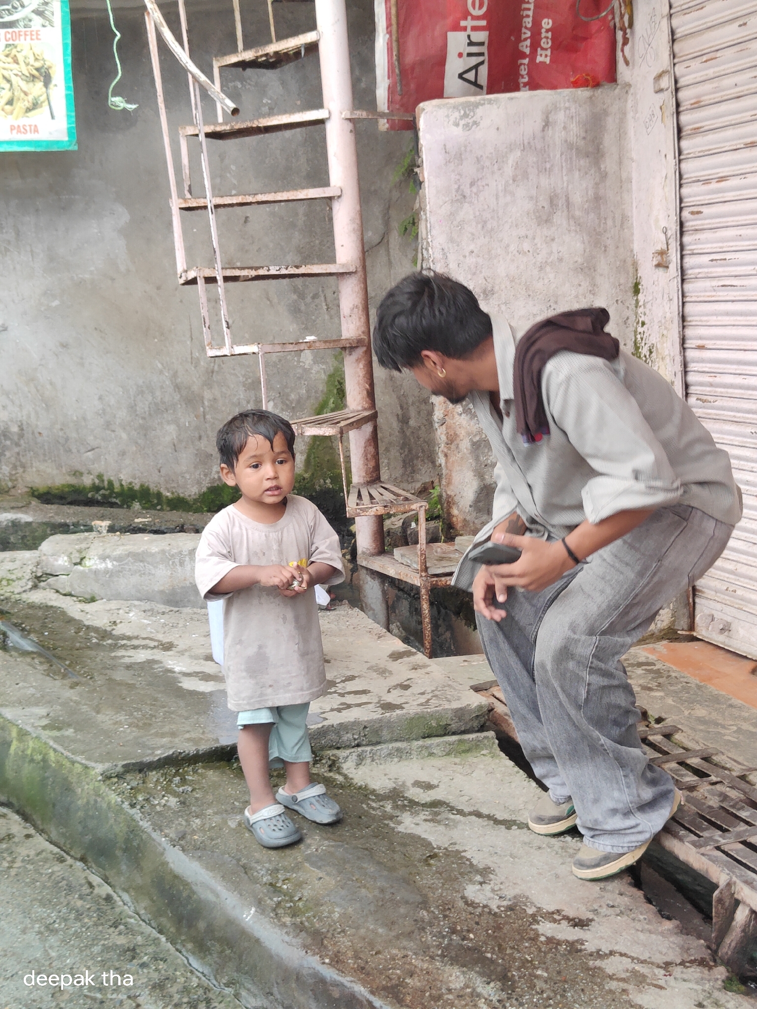 A boy and an adult man are standing on a wet concrete pathway in what appears to be an outdoor alleyway in Vashisht, India. The boy, on the left, is young, wearing a light-colored t-shirt and light blue shorts with gray clogs. He is holding some small objects in his hands and looking towards the man. The man, on the right, is wearing a light-colored button-up shirt and gray jeans. He has a dark cloth draped over his shoulders and is holding a dark object in his right hand, possibly a mobile phone, and is looking down. Behind them is a spiral staircase made of metal with a pinkish-brown hue, set against a textured concrete wall. To the left of the boy is a sign with text, and a green string is hanging from a railing above. The ground is damp with visible water channels and some green moss on the concrete structures. A metal grate is visible in the foreground to the right.