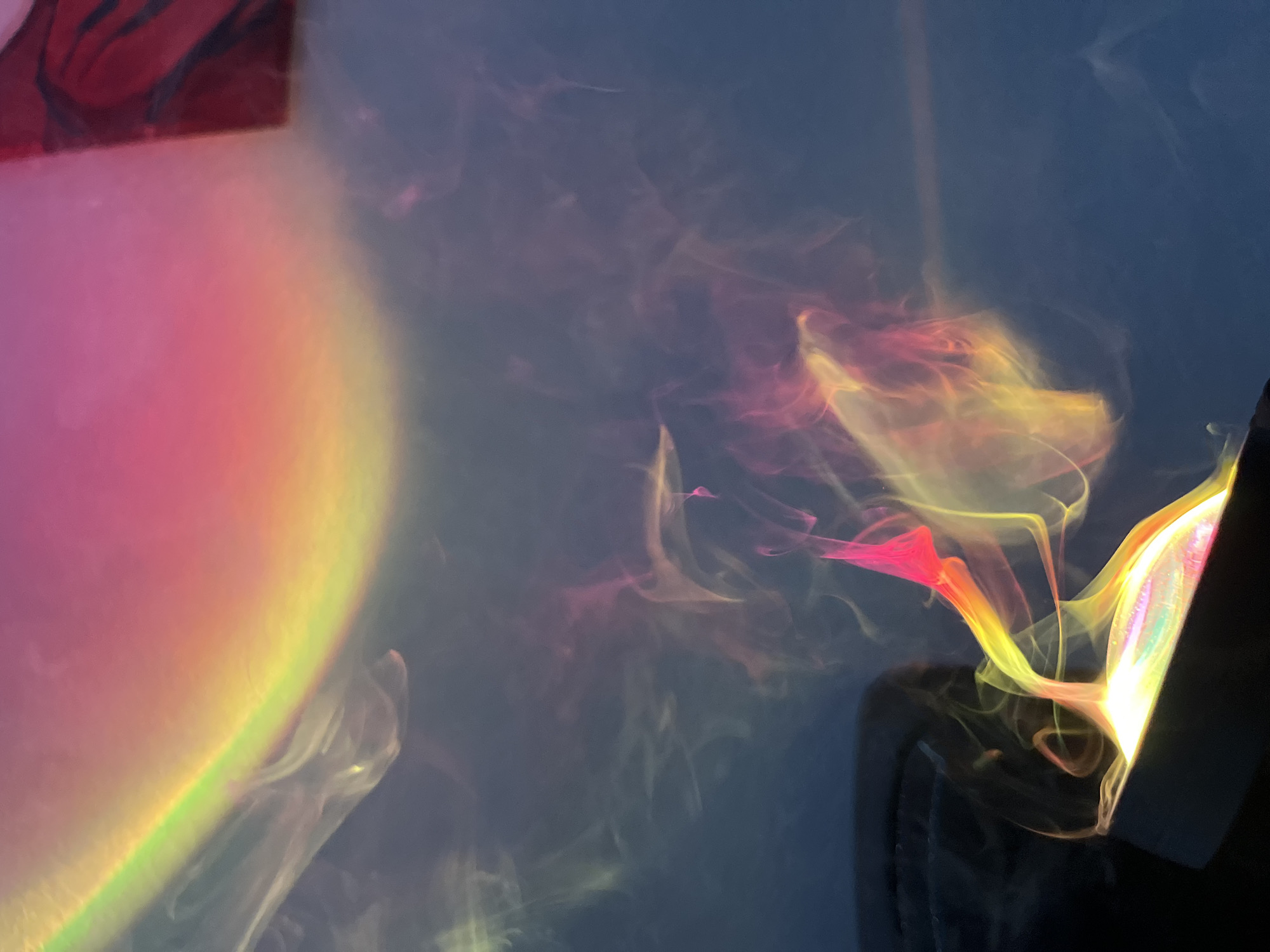 The image shows light and shadows. A multicolored light source is visible in the bottom left and right corner of the image. A black object is located in the bottom right corner. The background appears to be a wall, and the colors create a soft, ethereal effect. The specific location in the city of Unknown, Unknown is unknown.
