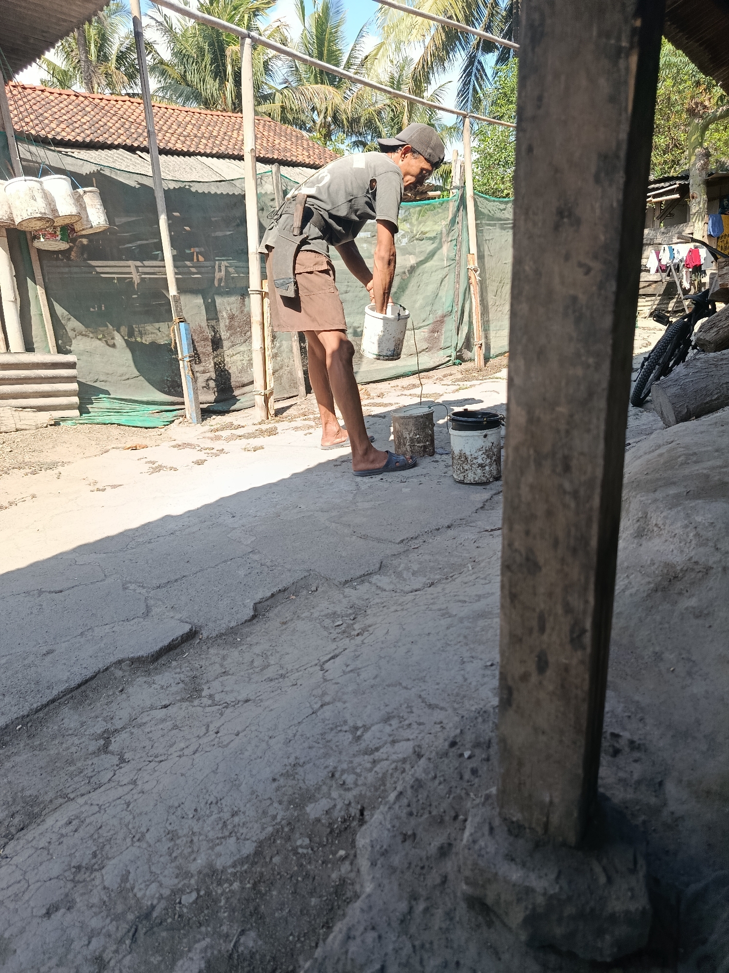 In Sarwadadi, Indonesia, a man in a dark t-shirt and brown shorts is seen working outdoors. He is wearing a dark baseball cap and flip-flops. The man is bent over, holding a white bucket in his hands as if about to pour its contents into another bucket on the ground. There are three buckets visible in the scene: one the man is holding, and two on the ground near him. The ground is a rough, cracked concrete surface, and a large shadow stretches across it from the right side of the frame. Behind the man, a tiled roof structure is visible, with palm trees and a blue sky overhead. Various objects, including what appear to be corrugated metal sheets and green tarpaulin, are present in the background, along with a bicycle and some laundry hanging on a rack. The overall scene depicts a person engaged in manual labor in a somewhat rustic outdoor setting.