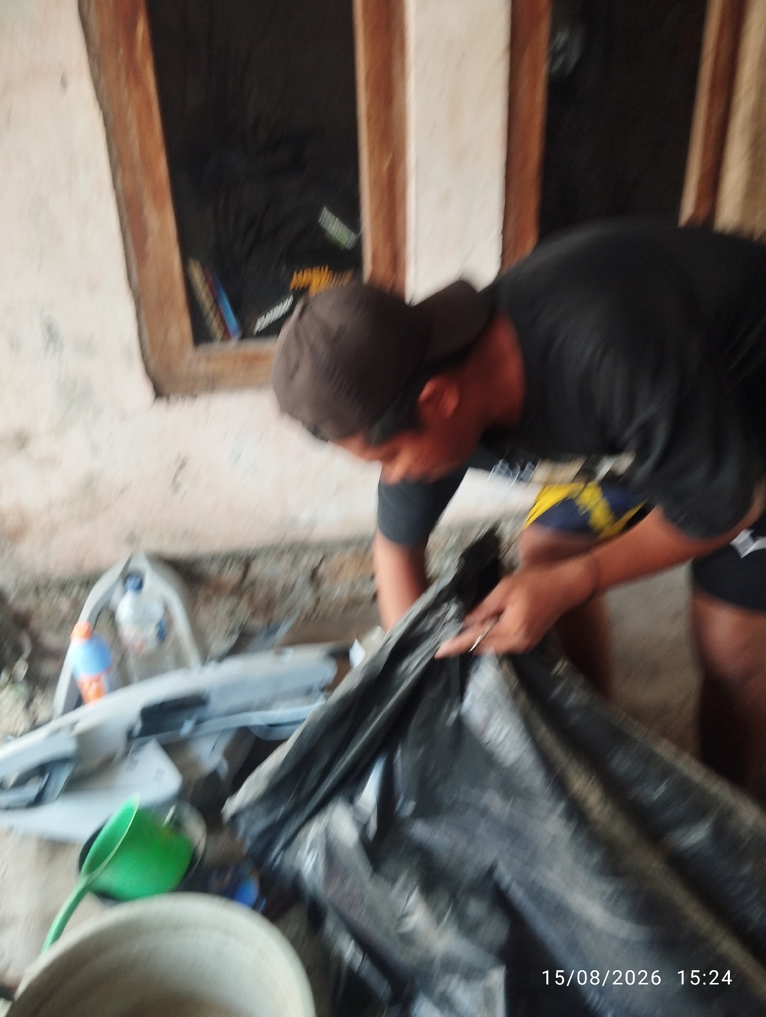 An outdoor scene in Tegal Rejo, Indonesia, shows a person in a dark t-shirt and a brown baseball cap bending over a large black plastic bag. The person is wearing yellow and blue patterned shorts. Their hands are engaged with the bag, suggesting an action of gathering or securing its contents. In the background, a pale, textured wall with two wooden-framed openings is visible. Scattered on the ground are various objects, including a grey plastic container with a blue bottle inside, an orange and blue bottle, and a green scoop. The overall environment appears to be somewhat cluttered and possibly under renovation or storage. The timestamp on the image reads "15/08/2026 15:24".