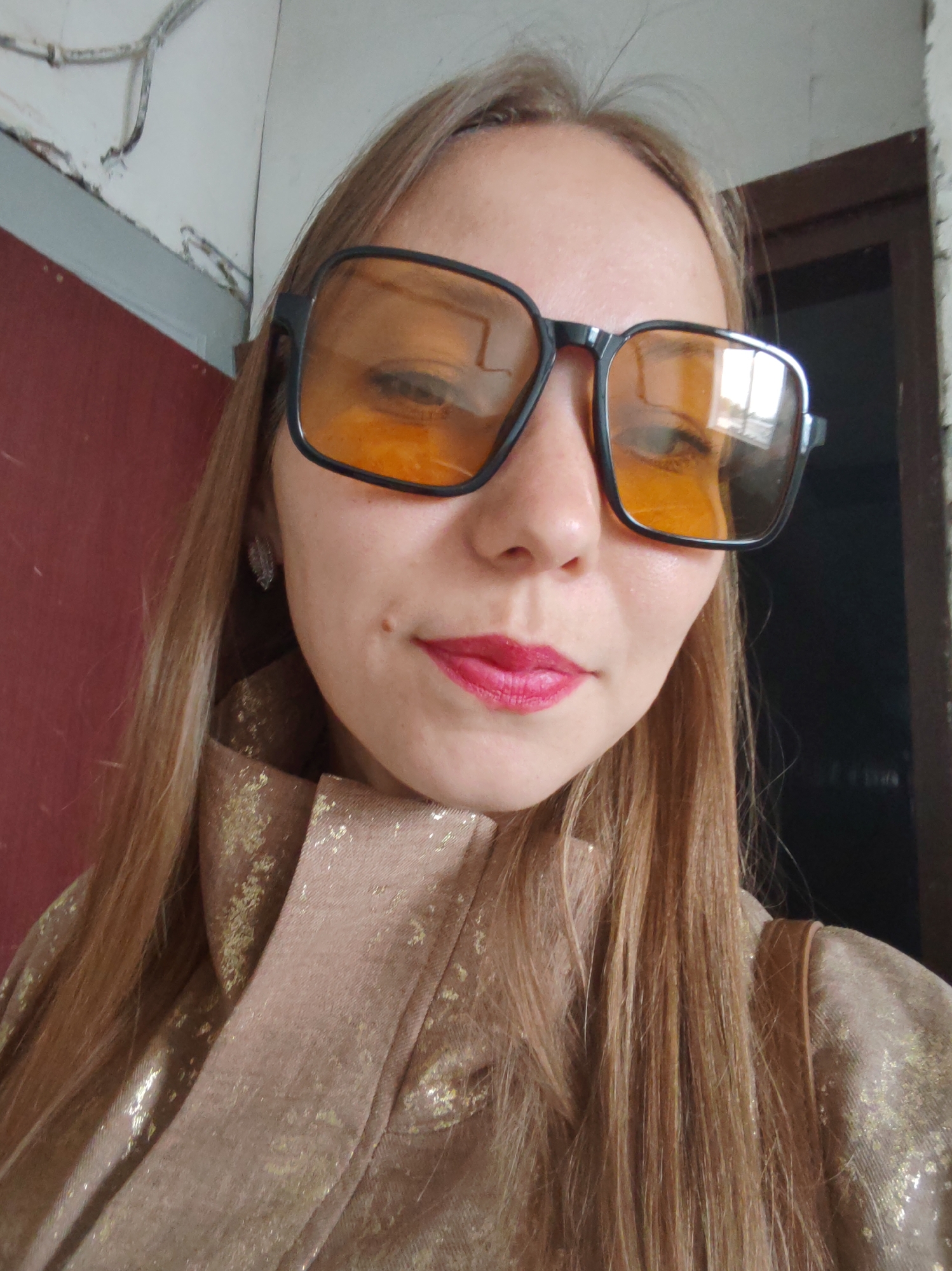 The image is a close-up, selfie-style shot of a young Caucasian woman with blonde hair, wearing oversized black-framed sunglasses with yellow lenses and bright pink lipstick. She is wearing a tan jacket with a shimmering gold pattern. She appears to be indoors, with a reddish wall on the left and a dark doorway or opening on the right. The overall environment suggests an urban setting, potentially an apartment or a less-maintained public space, but there is no specific visual information to confirm the location as Syktyvkar, Russia. The lighting is soft and diffuse.
