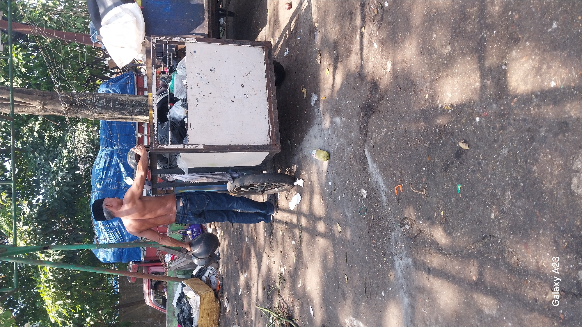 A shirtless man wearing a black peci stands in an outdoor, cluttered environment. He is holding a dark-colored helmet in his left hand, and his right hand rests on the edge of a white, metal-framed cart filled with trash bags. The cart has a single wheel visible. In the background, a truck covered with a blue tarp is partially visible. The ground is paved and appears dirty, with scattered debris, including a yellow plastic bottle. Trees and foliage are present behind the man and the truck. The scene appears to be in an urban setting, possibly a roadside or an open market area in Malang, Indonesia, given the presence of the peci, common headwear in Indonesia.