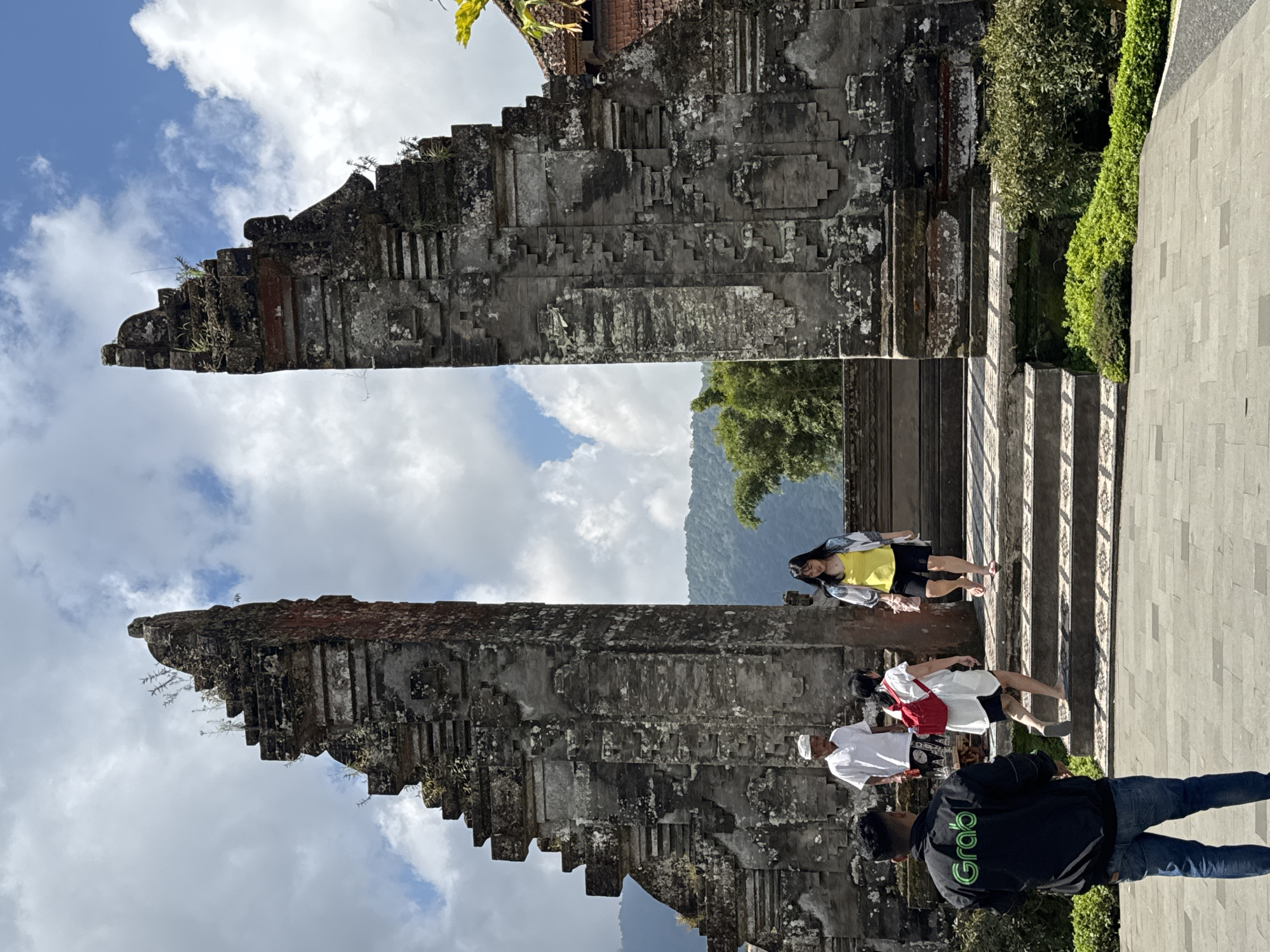 The image shows a group of people standing on steps in front of a large stone gate. The gate is made of gray stone and has a tiered, angular design. The sky is visible through the gate's opening. The location is in Baturiti, Indonesia. There are four people visible. One person wears a black jacket with "Grab" printed on it. A woman in a yellow shirt stands near the top of the steps.
