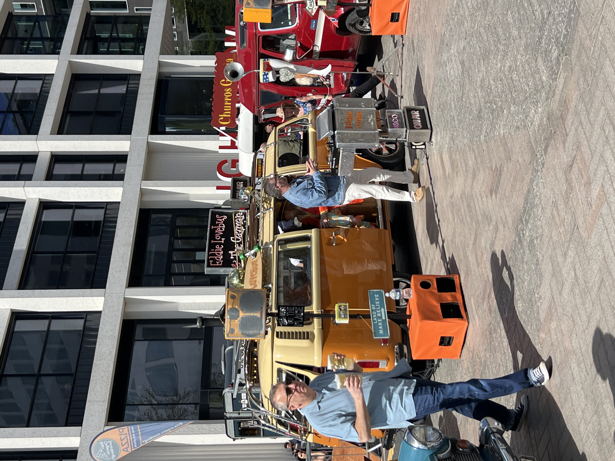 The image shows a scene featuring a gathering of decorated vehicles, possibly for an event or performance, set against a modern architectural backdrop, possibly in London.
**People:** Two men are prominently visible, standing near or interacting with the vehicles. The first man, dressed in a light blue shirt and jeans, holds what appears to be two beer glasses and has sunglasses. The second man is wearing a denim jacket and white pants. Other people can be seen in the vehicles.
**Subjects/Objects:** The main subjects are the decorated vehicles. A brightly colored, possibly vintage, van with speakers and signs like "Eddie Lovebus" and "Land of Make Believe" is prominent. Another red vehicle with a churros sign is positioned behind the yellow van. There is a scooter. Speakers, signage, and various decorative items are mounted on the vehicles.
**Actions/Interactions:** The men are standing and interacting with the vehicles, possibly as performers or attendees.
**Environment:** The vehicles are parked on a paved area, likely a street or plaza. A modern building with distinctive windows forms the background. The scene is captured during the day under bright, sunny conditions.
The presence of the 'Churros G' sign hints at the presence of a food stall nearby, and the vehicles are used for a celebration.