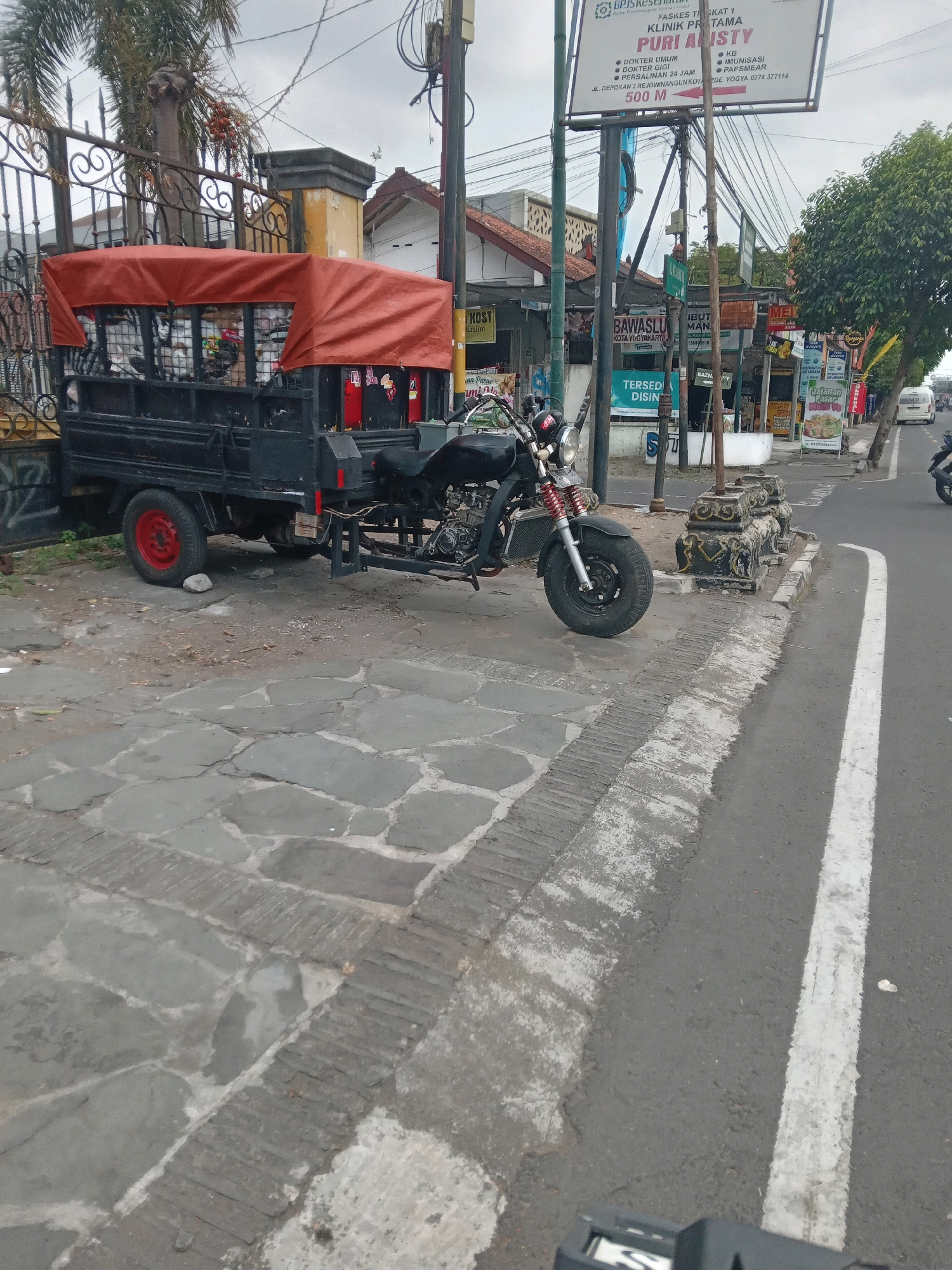The image depicts a street scene in Yogyakarta, Indonesia, featuring a three-wheeled vehicle, possibly a motorcycle with a cargo bed, parked on a paved area next to a road. The vehicle has a black cargo bed covered with an orange tarp, and a motorcycle engine and front wheel are visible. The ground is paved with large, irregular stone slabs, with a textured concrete border separating it from the asphalt road. A white line marks the edge of the road. In the background, there are buildings, trees, and a signpost with multiple advertisements and informational signs, including one for "PURI ALISTY" and "BAWASLU KOTA YOGYAKARTA." A black metal fence is visible to the left of the vehicle. The sky is overcast. No people are clearly visible in the image.