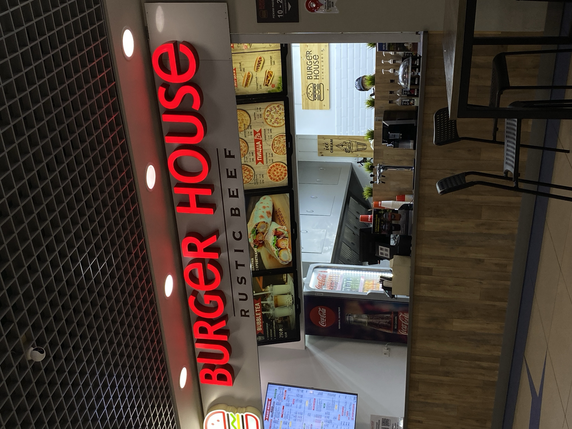 The image shows a Burger House fast-food restaurant in Votkinsk, Russia. The restaurant features a prominent red neon sign that reads "BURGER HOUSE" and "RUSTIC BEEF." The interior is lit with bright lights, and the counter area displays menu boards with food items like burgers, pizzas, and bubble tea. A Coca-Cola refrigerator is visible behind the counter. There are several chairs and tables in the foreground, suggesting a dining area. A security camera is mounted on the ceiling. The overall scene depicts a typical fast-food establishment.