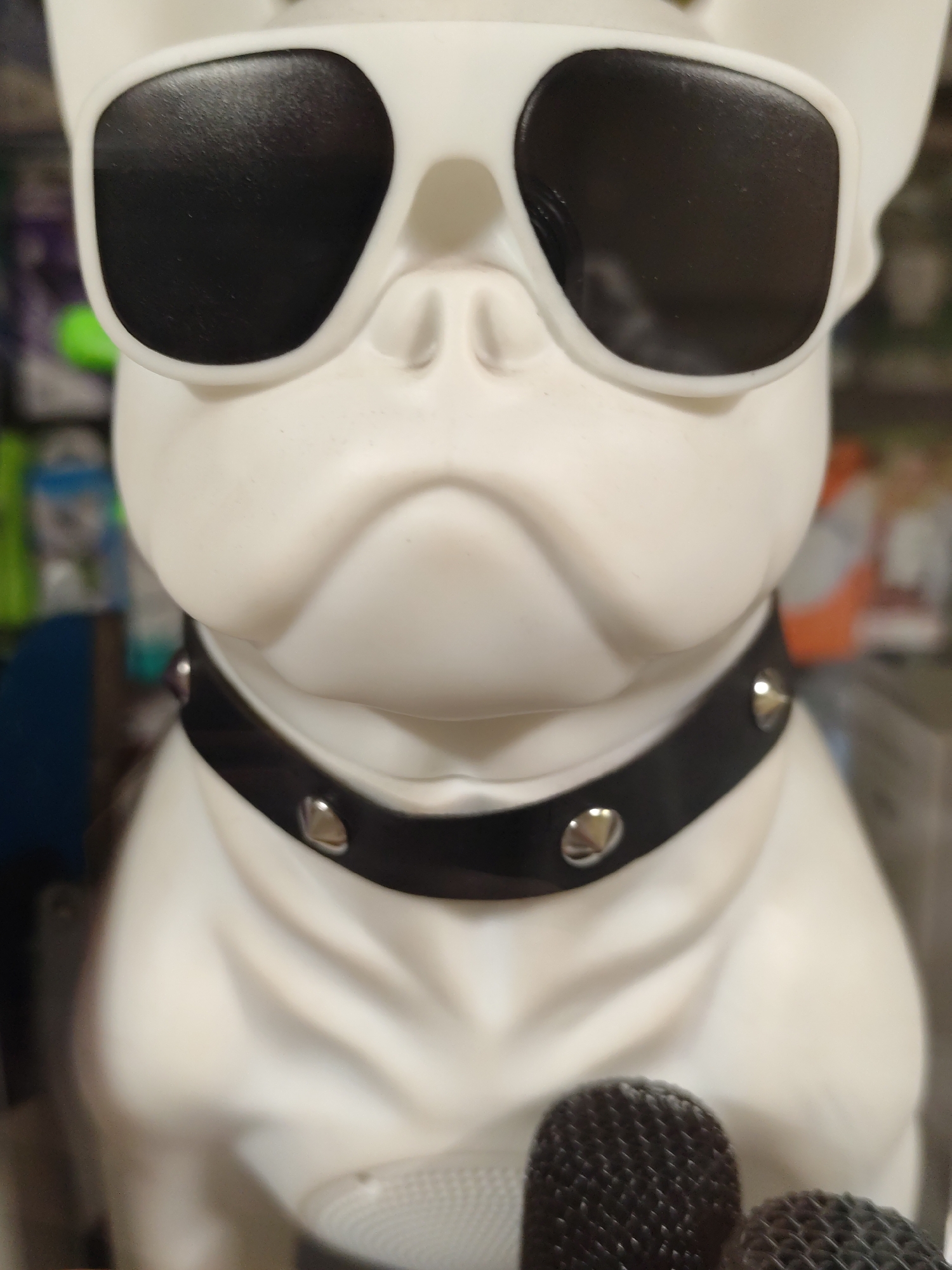 This is a close-up, eye-level, medium shot of a white, stylized dog figurine, possibly a French bulldog, wearing black sunglasses and a black studded collar. The figurine is positioned centrally and takes up most of the frame. The background is out of focus and appears to be an indoor setting, with hints of shelves and various objects, suggesting a retail environment. The lighting is artificial and illuminates the figurine from the front, creating some shadow on the left side of its face. There are two microphone heads visible in the foreground, partially obscuring the bottom of the figurine. No location in Moscow, Russia, is identifiable from the image.