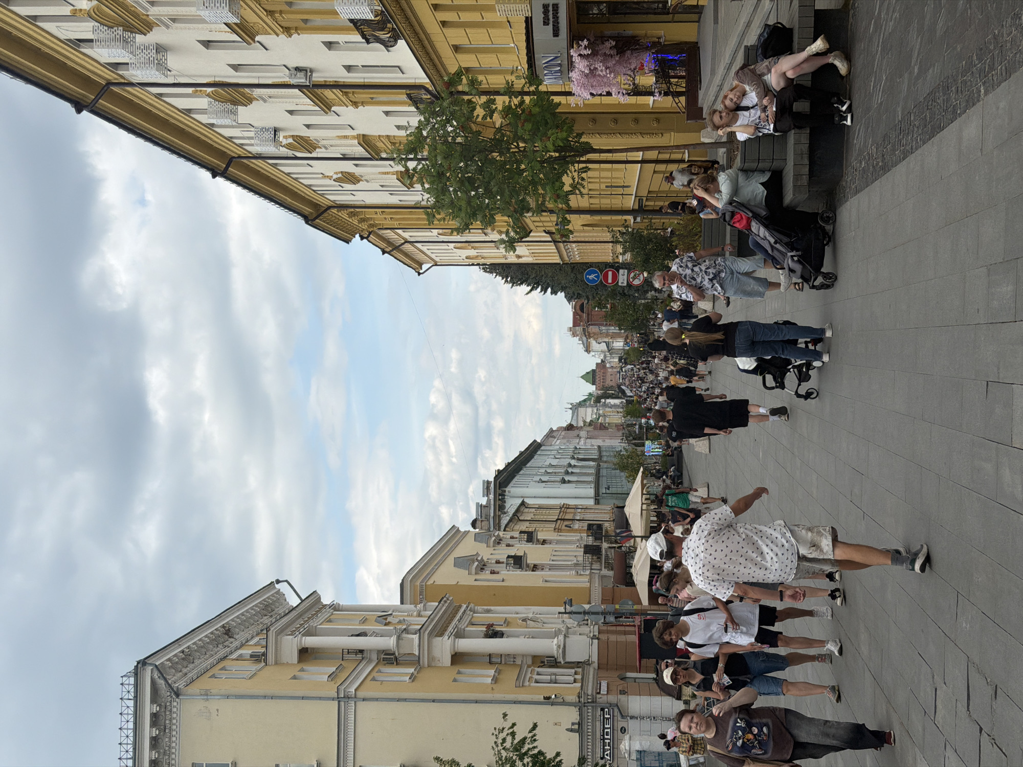 The image depicts a busy pedestrian street in Nizhny Novgorod, Russia. The street is lined with tall, ornate buildings, primarily yellow and white, with architectural details suggesting historical significance. The sky is overcast with scattered clouds.
On the street, a diverse crowd of people are walking, with some gathered in small groups. Many are dressed casually in summer clothing. In the foreground, several individuals are walking towards the viewer, including a woman in a dark t-shirt and jeans, a man in a patterned white short-sleeved shirt and shorts, and a young man in a white t-shirt and black shorts.
Further down the street, more people are visible, some pushing strollers. A couple is seated on a bench on the right side of the image, with the woman leaning her head on the man's shoulder. Shops and cafes are visible along the sides of the street, with awnings providing shade. Signs in Russian are present, indicating businesses and street information.
The overall atmosphere is one of a lively, urban public space, likely a popular area for shopping and recreation in the city. The architecture and street layout suggest a central European or Russian urban environment.