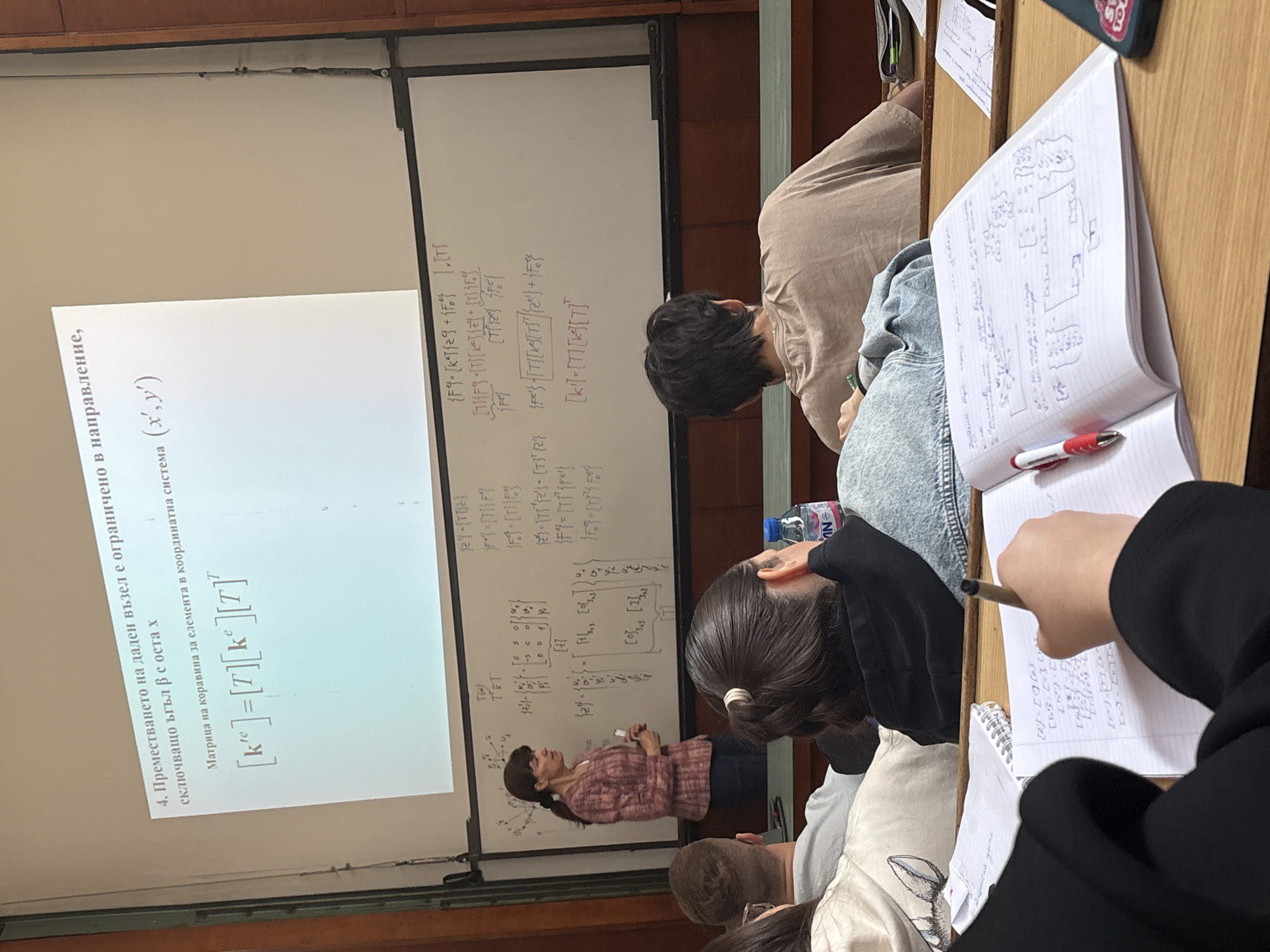 The image shows a classroom setting, likely in Sofia, Bulgaria, given the Cyrillic script on the projector screen. A female professor is presenting a lecture, with equations and text displayed on the screen behind her. Several students are seated at desks, taking notes in notebooks. The notes include handwritten equations and diagrams, suggesting the class is related to mathematics, physics, or engineering. The room is well-lit with a whiteboard, projector, and rows of desks and chairs. A bottle of water is visible near a student, and various personal items are scattered on the desks.