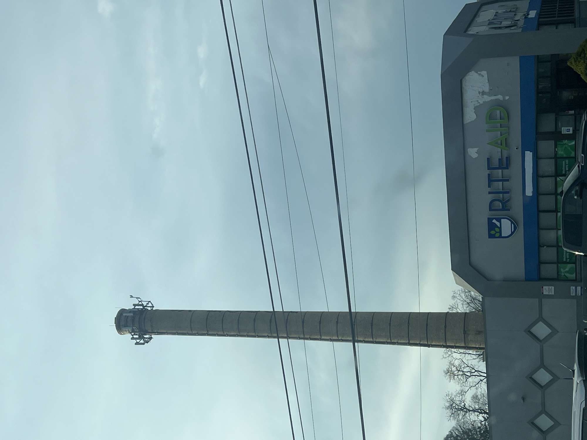 The image depicts a tall, brick smokestack in the City of Newburgh, New York. In the foreground, there is a Rite Aid store. Power lines cross the frame. The sky is overcast. Bare tree branches are visible near the base of the stack.