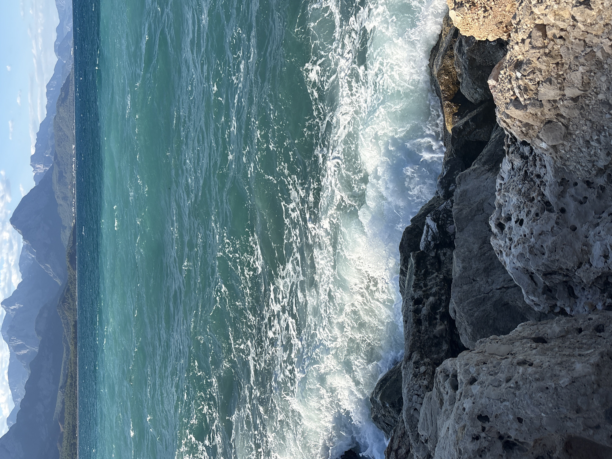 A wide-angle, vertical, eye-level shot captures a coastal scene with rough waves crashing against a rocky shore in Kemer, Turkey. The ocean, a vibrant turquoise, occupies the majority of the frame, with white foam cresting as waves break against the boulders. In the distance, a range of majestic mountains, some with snow-capped peaks, form a dramatic backdrop against a hazy blue sky dotted with white clouds. The foreground is dominated by large, textured rocks, some appearing porous and weathered, others darker and more jagged. The lighting suggests it is either early morning or late afternoon, casting a soft glow on the landscape. No people or animals are visible in the image.