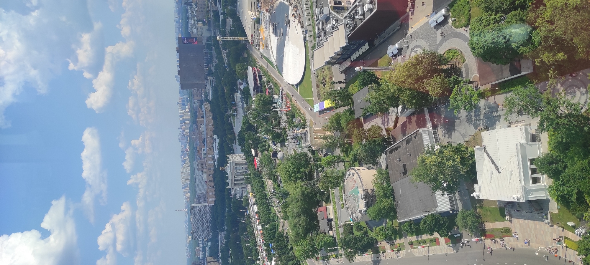 The image is an aerial view of a park and urban area. In the foreground, there are several buildings and a white, multi-gabled structure. A street with pedestrians is visible on the left. Further back, a circular building with graffiti is surrounded by trees. Beyond this, the park features green spaces, trees, and what appear to be amusement park rides and structures. A large, ornate building with a dome and columns is prominent in the middle ground. In the background, a dense urban landscape with numerous buildings, including high-rises, stretches to the horizon. The sky is blue with scattered white clouds. The scene appears to be set in the Ostankino district of Moscow, Russia, given the architectural style and the presence of what resembles the VDNKh park.