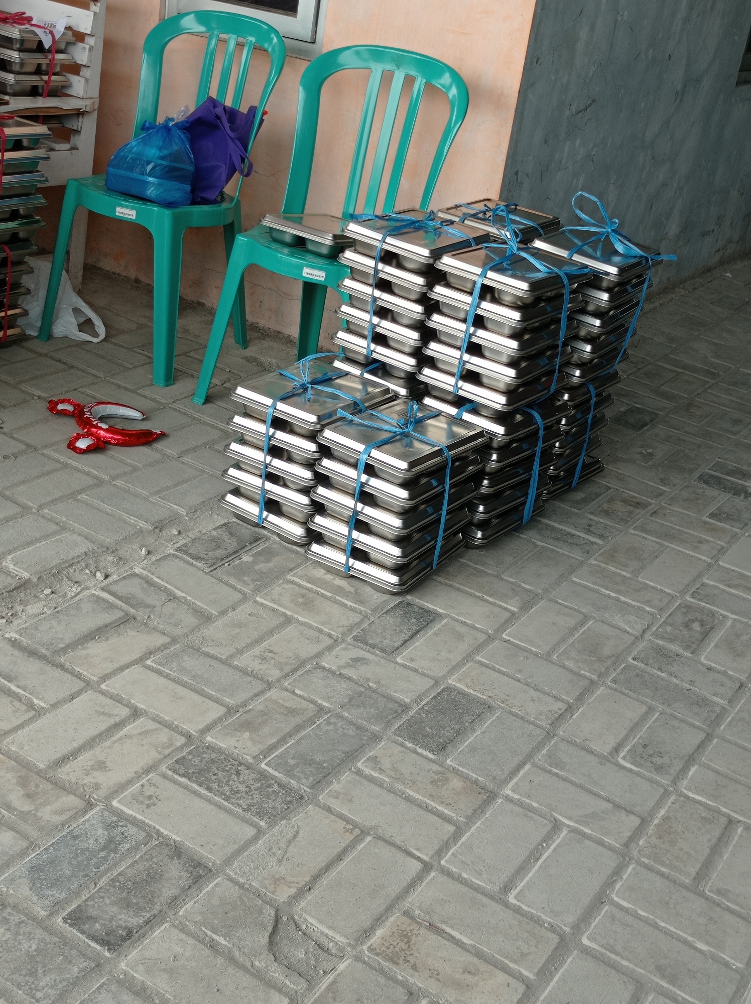 In the city of Maribaya, Indonesia, this image captures an outdoor scene on a paved walkway. Two teal plastic chairs are placed side by side. On the left chair, a blue plastic bag and a dark purple bag rest. To the right of the chairs, stacks of rectangular metal containers, possibly food trays, are neatly piled. These stacks are secured with blue string. In the foreground, a deflated red balloon, shaped like a pair of lips, lies on the ground. The background features a textured wall, appearing to be painted in shades of peach and grey. The paving stones are arranged in a pattern.