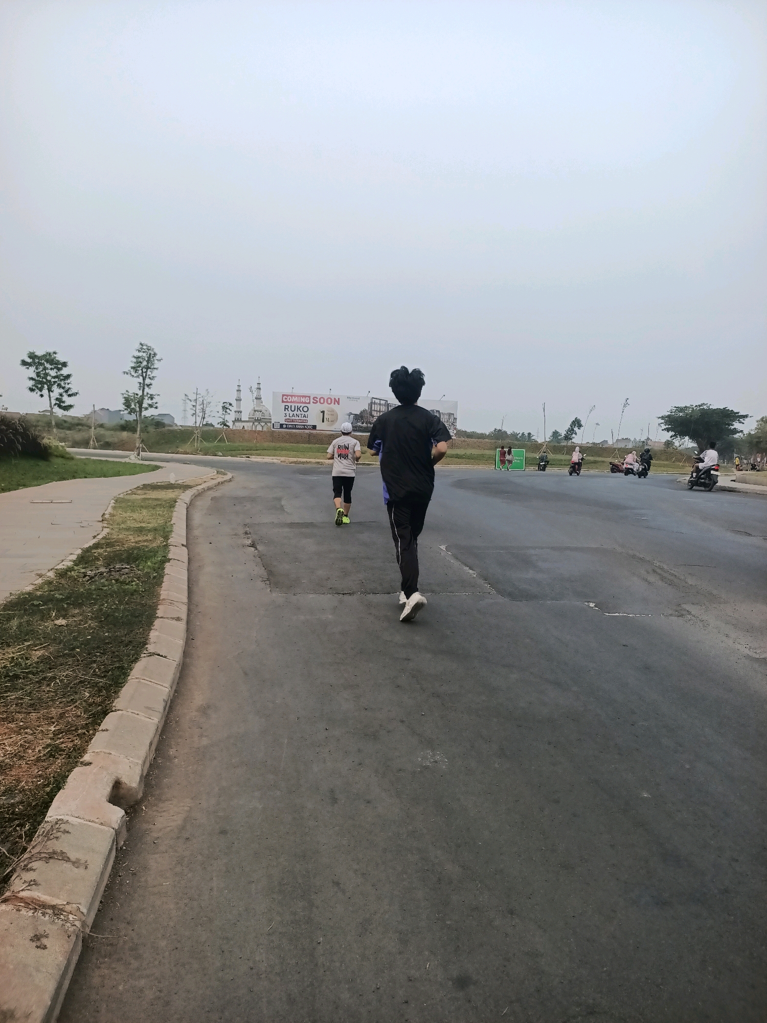 The image shows two individuals running on a paved road in Telagamurni, Indonesia. The person in the foreground is wearing a black shirt and black pants, with white sneakers. The person behind them is wearing a white t-shirt with red lettering and dark shorts, also with white sneakers and a white cap. They are running away from the camera. The road appears to be a street or pathway within a development, with a sidewalk and grass to the left. In the background, there are trees, a billboard advertising "COMING SOON RUKO 3 LANTAI", and several motorcycles parked or moving on the road. The sky is overcast and the overall lighting suggests it is either early morning or late afternoon.